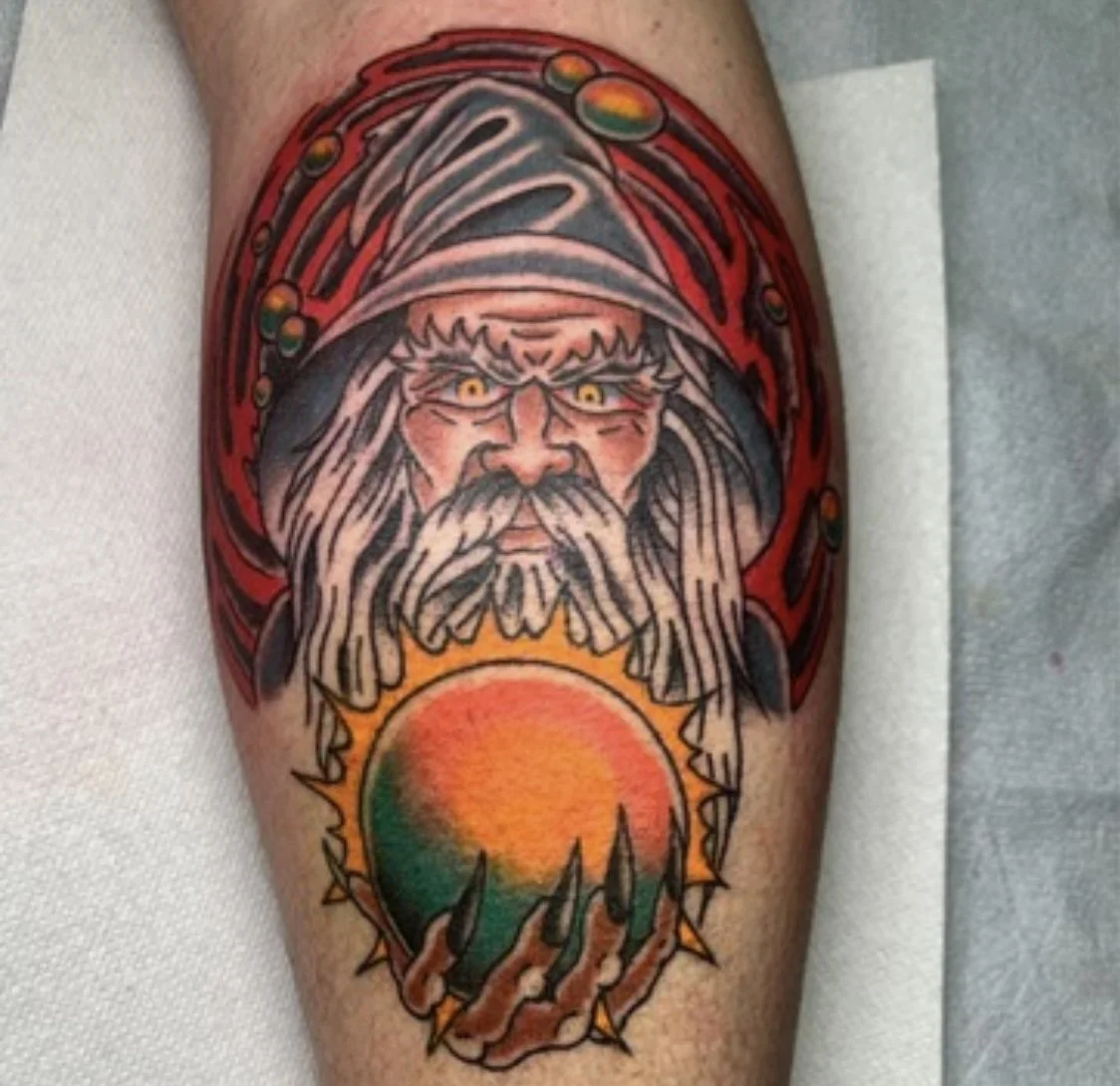 Tattoo of a wizard with a long beard and yellow eyes holding a sun with orange, yellow, and green colors, surrounded by red and black details. Traditional Tattoo Shop Wizards Lubbock,Texas 
Best of Lubbock Walk In tattoo Shop 