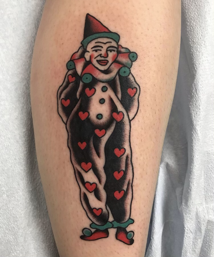 A tattoo of a person with a clown hat and makeup, dressed as a clown with a black and white body, decorated with red hearts, and wearing red shoes with green accents.