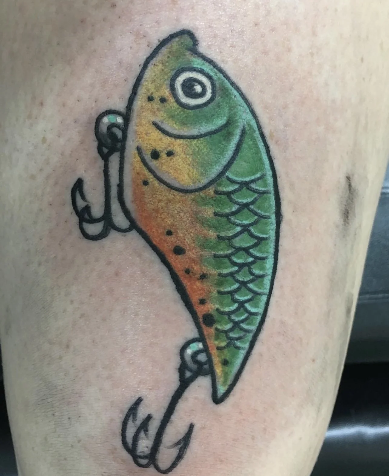 Colorful tattoo of a fish with green, orange, and yellow shading and black outlines on a person's skin.