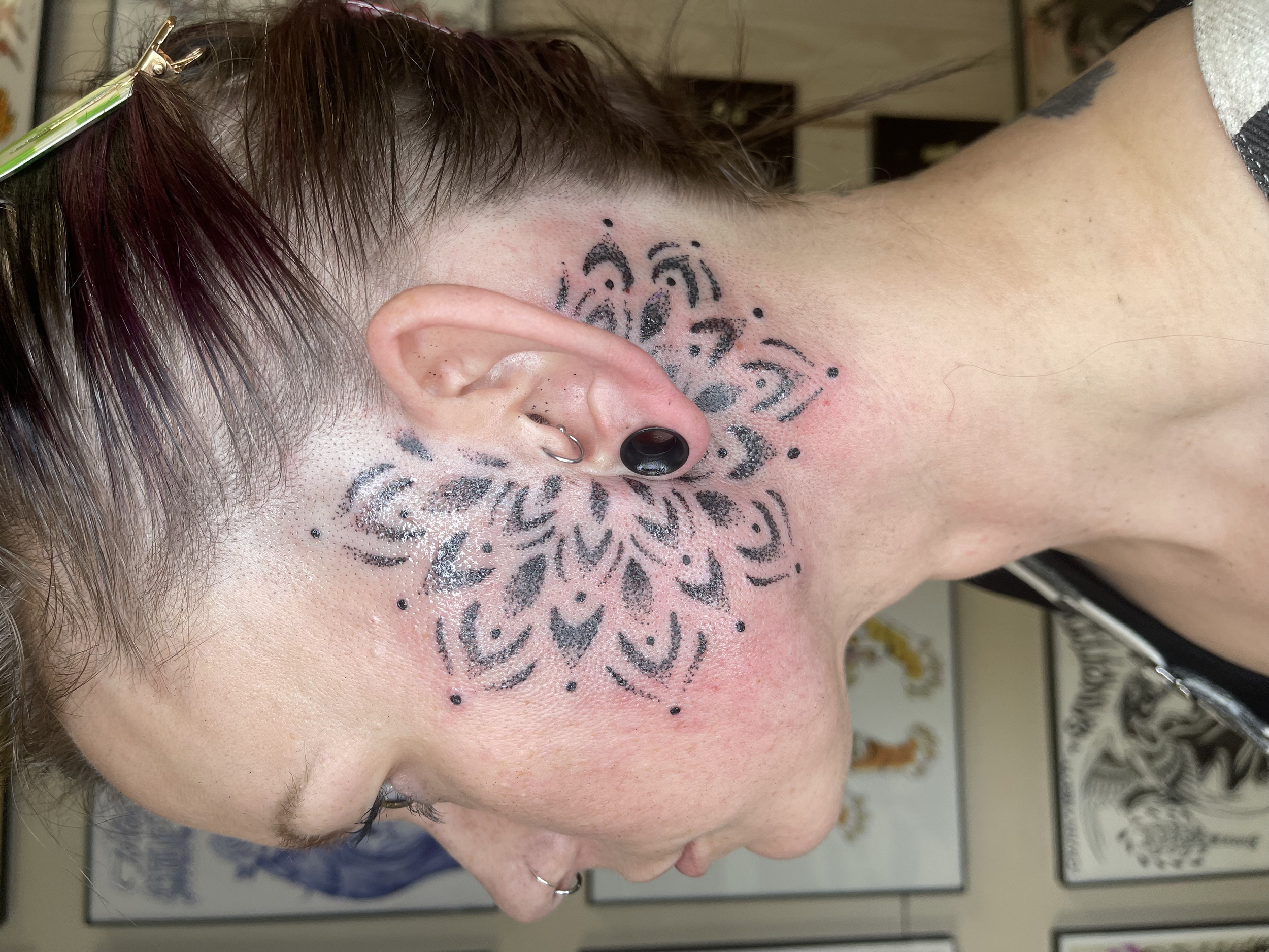 Side view of a person with a tattoo on the face, ear piercings, and short hair, in a tattoo or piercing studio.