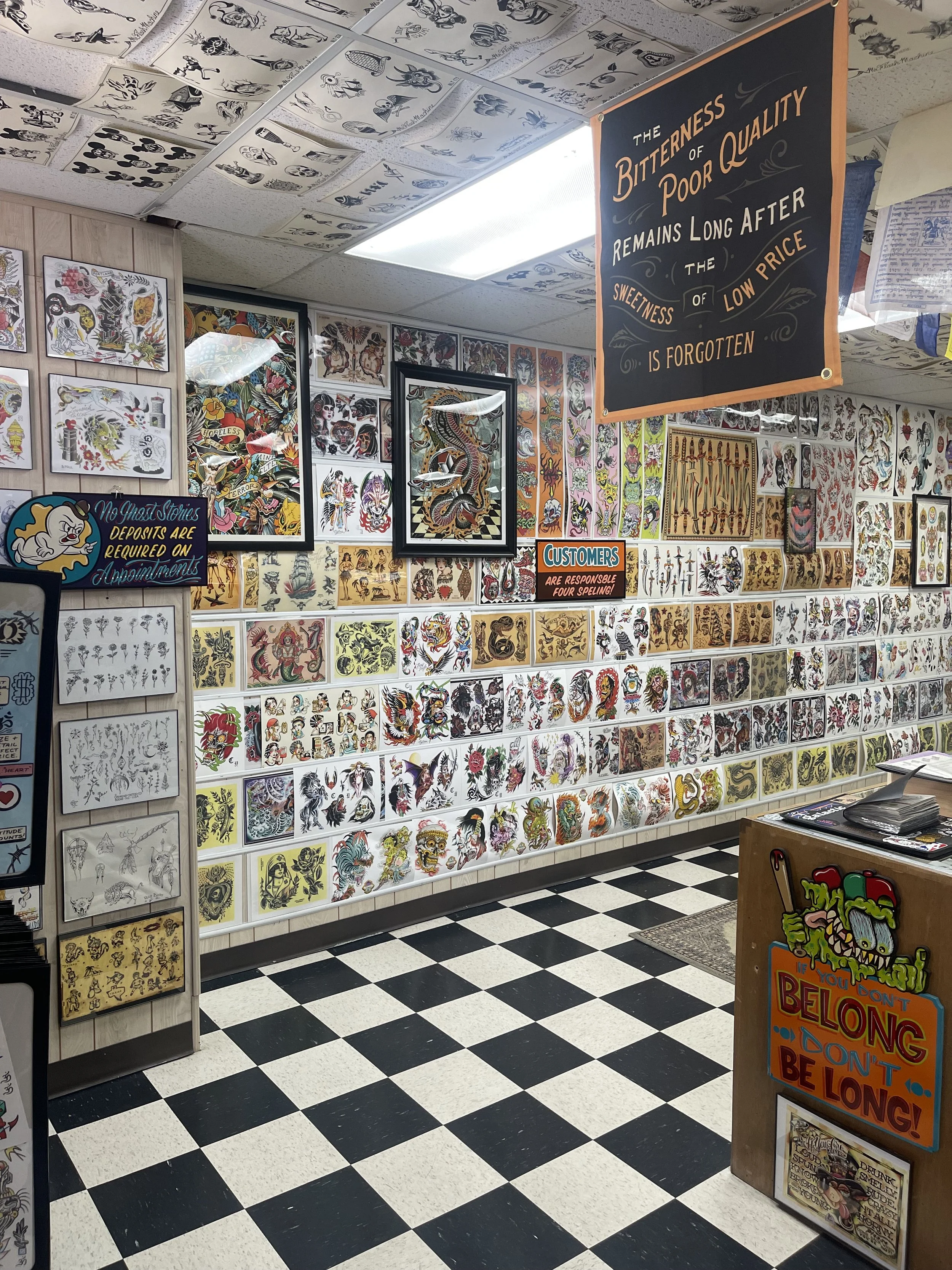 Tattoo shop interior showcasing walls covered with colorful traditional and modern tattoo flash art, including designs of skulls, dragons, butterflies, and historical imagery, with a sign on the wall about the influence of bad tattoos, and a small display table in the foreground with more tattoo artwork.