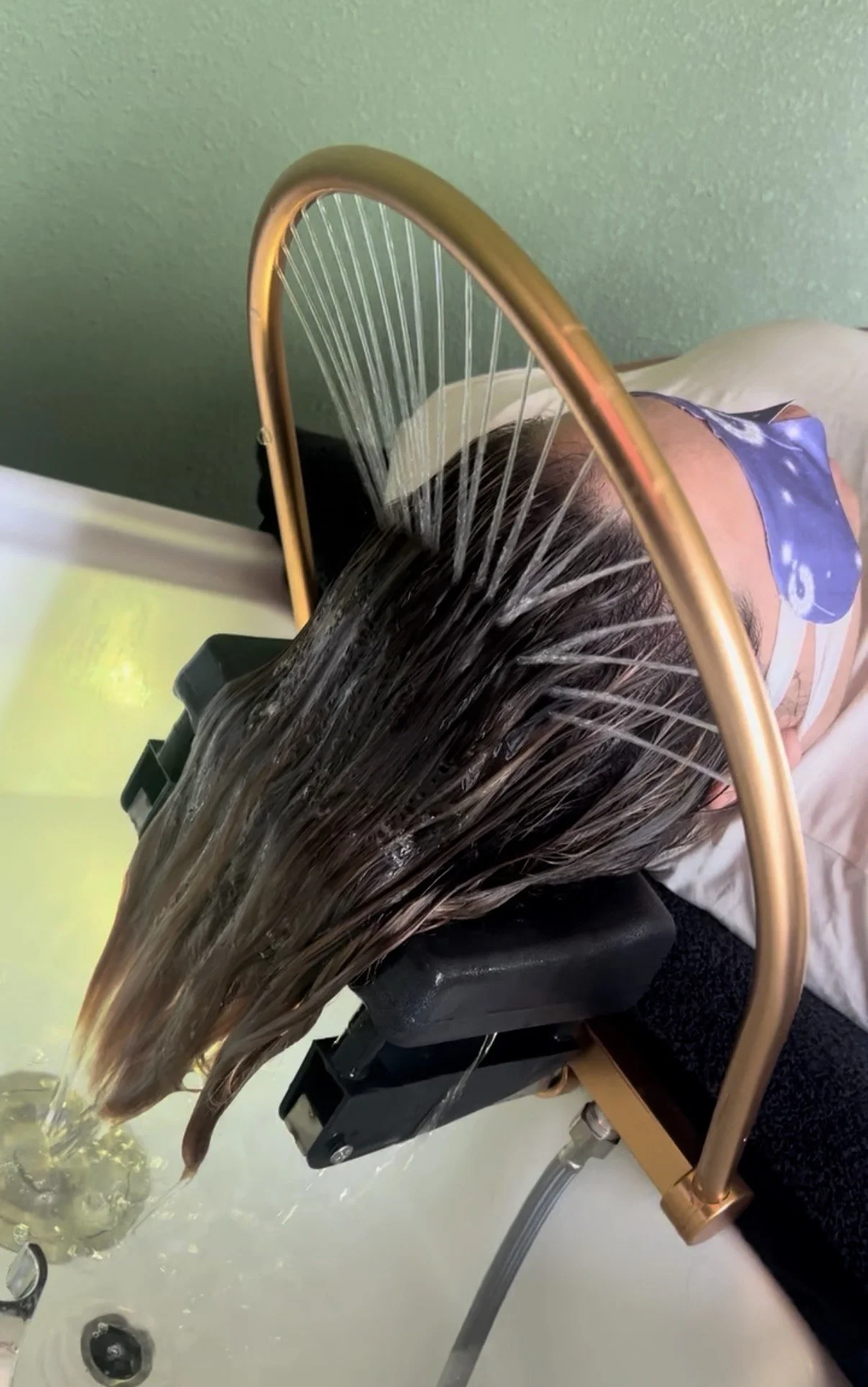 Person getting hair washed in a head spa bed with water flowing from the faucet, with a green wall in the background.