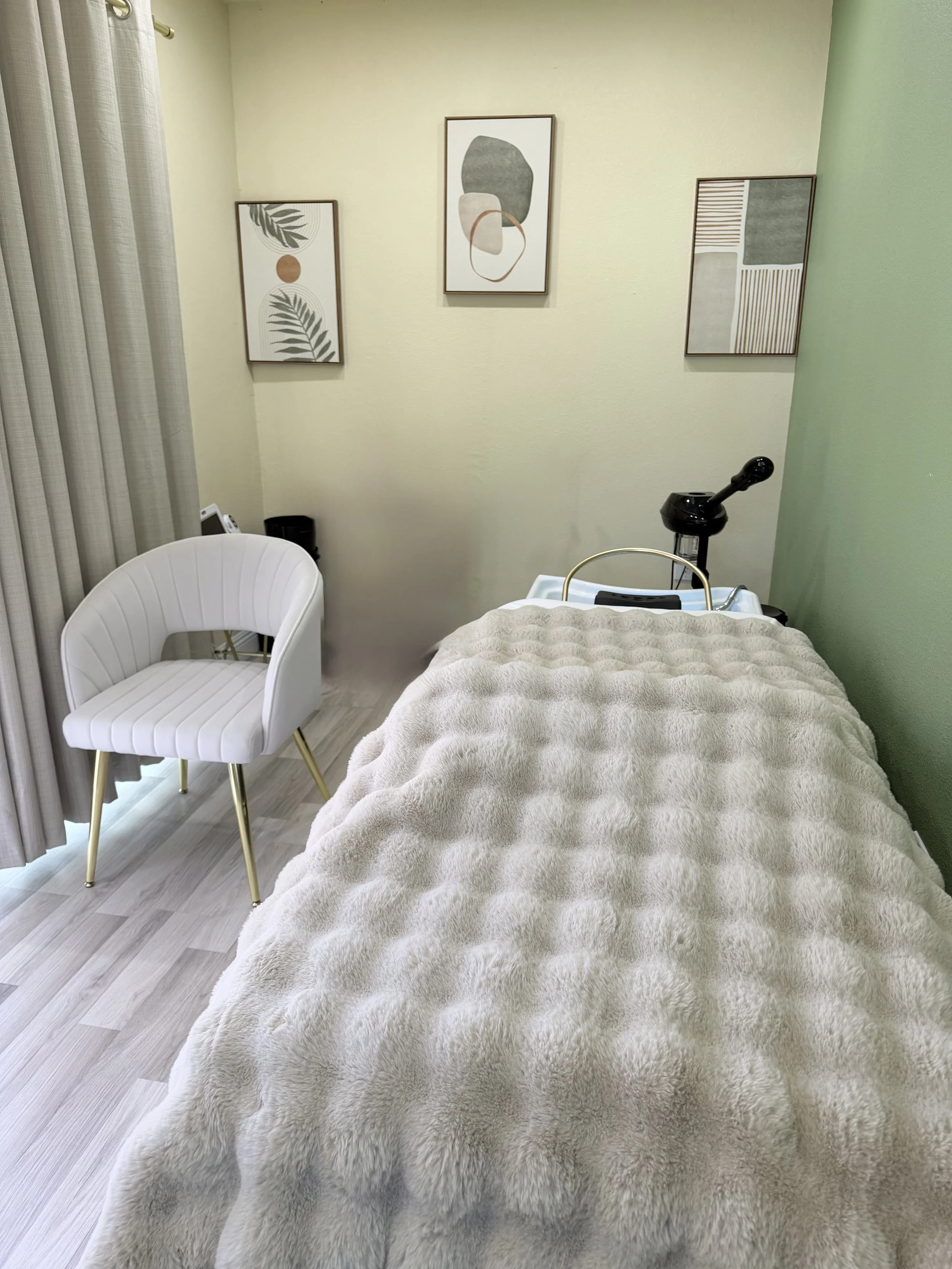 A head spa bed covered in a plush beige blanket, a white chair with gold legs, a black lamp, and three abstract framed pictures on a pale yellow wall. Light-colored wood flooring and beige curtains are also visible.
