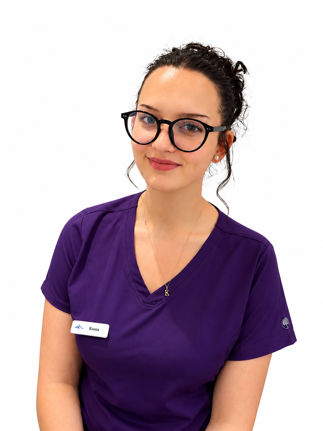 A young woman in purple scrubs with glasses, a name tag reading 'Ksenia,' and a gold necklace with a 'K' pendant, smiling at the camera against a white background.