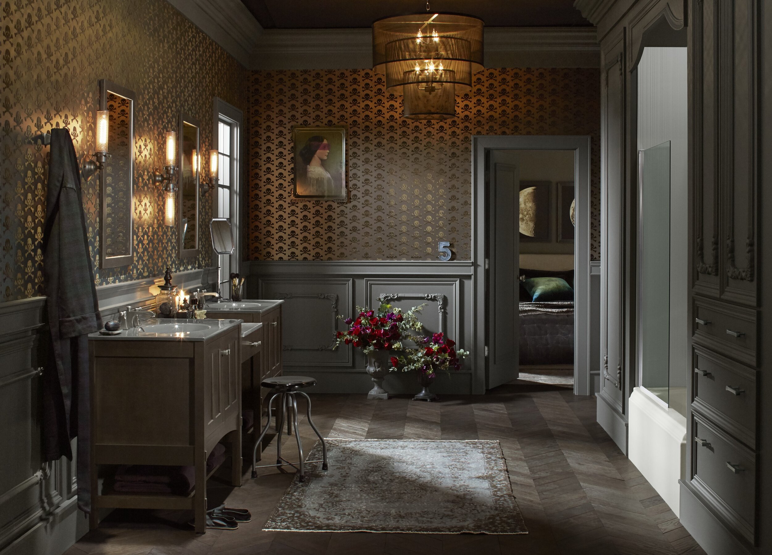 wide angle shot of bathroom with grey wainscot paneling underneath skulls velvet flock wallpaper with moon artwork and bedroom in the distant background