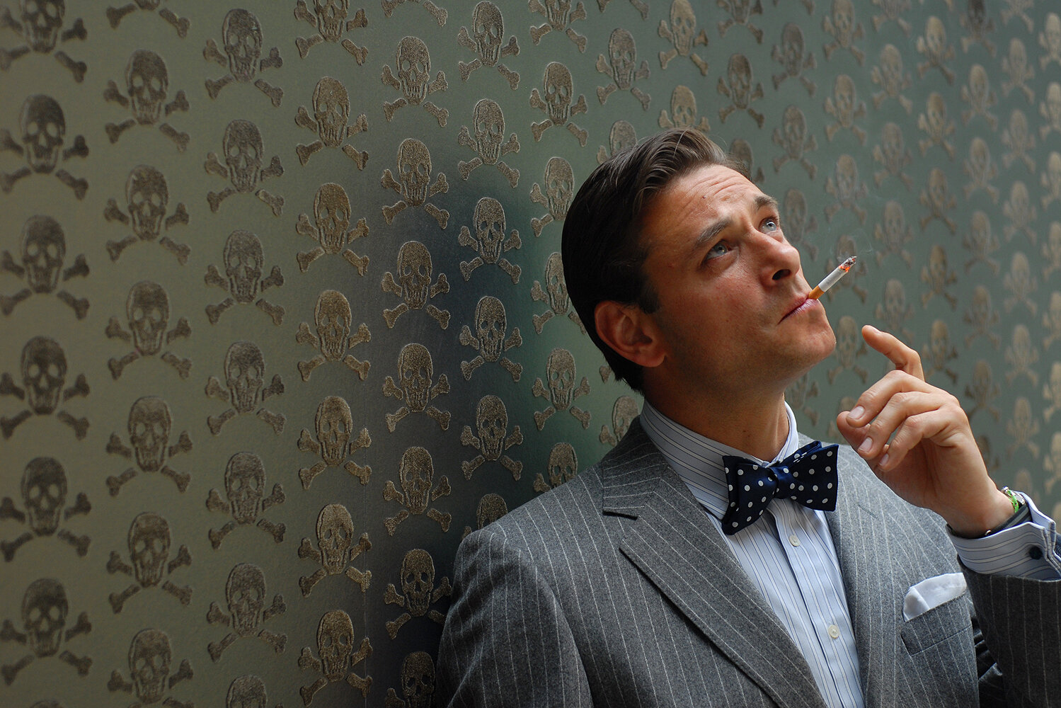 backing trader in bow tie and pin stripe suit smoking cigarette wistfully in front of skulls velvet wallpaper