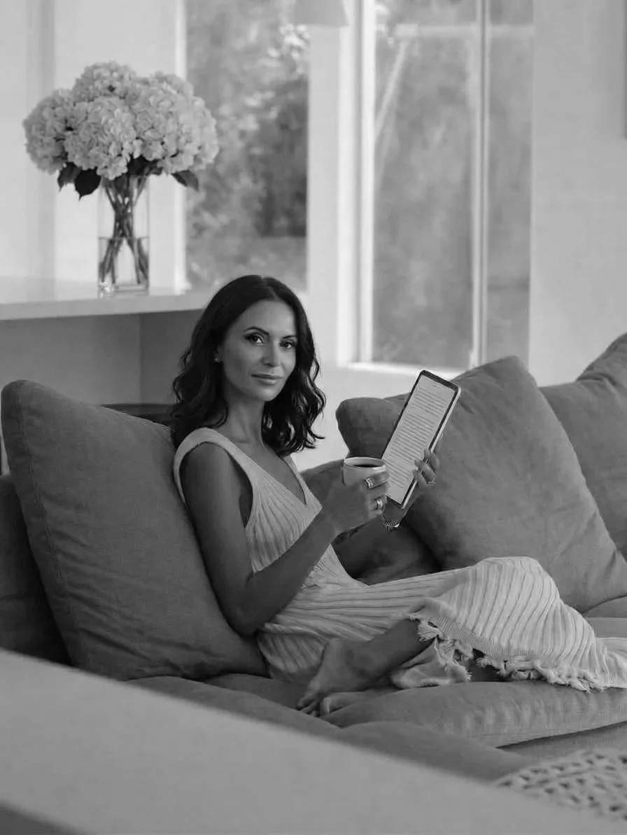 Black-and-white photograph of a woman seated on a sofa reading from a tablet.