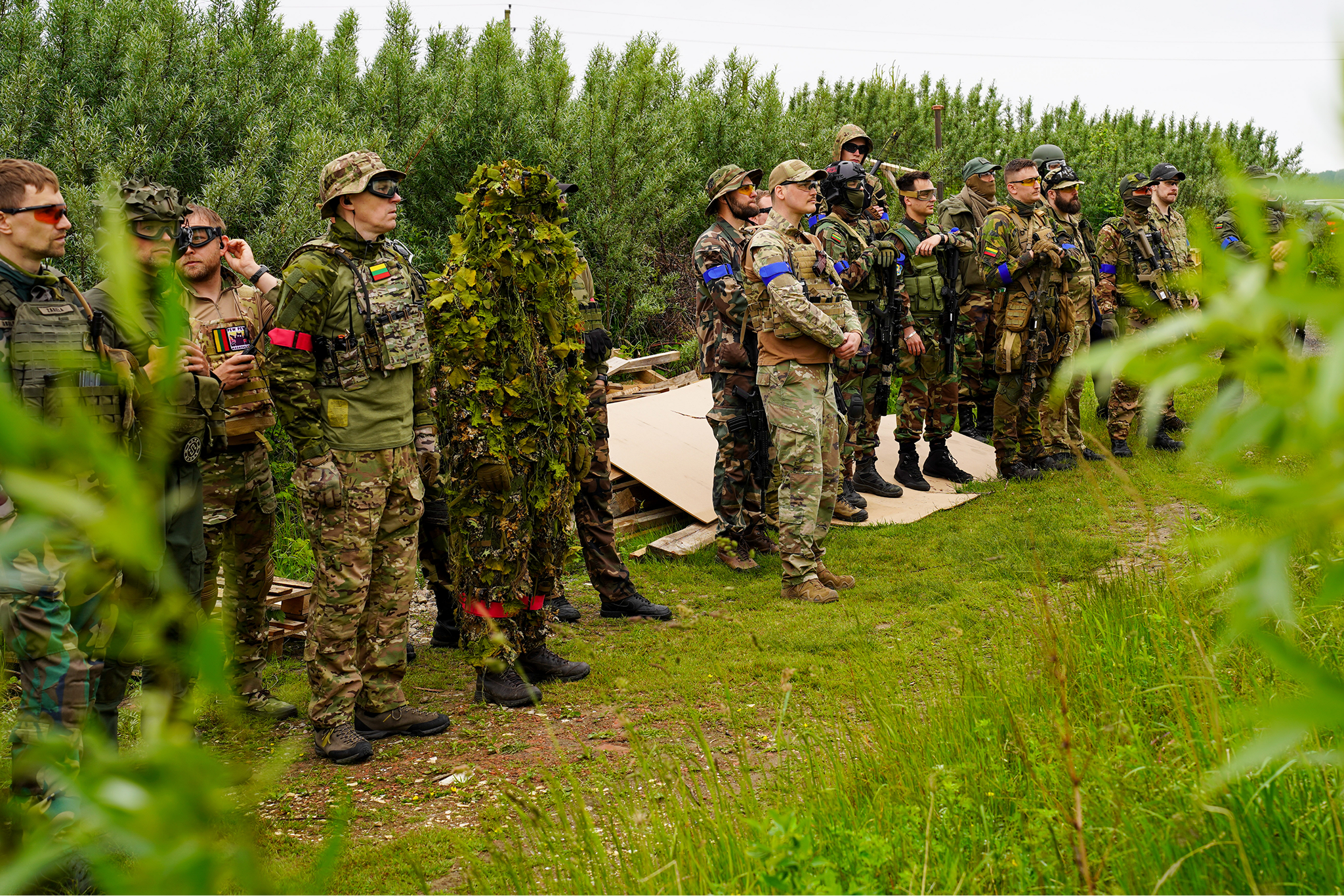 Group of soldiers in camouflage uniforms and tactical gear standing in a line outdoors on grass, with some wearing helmets and sunglasses, during daytime.