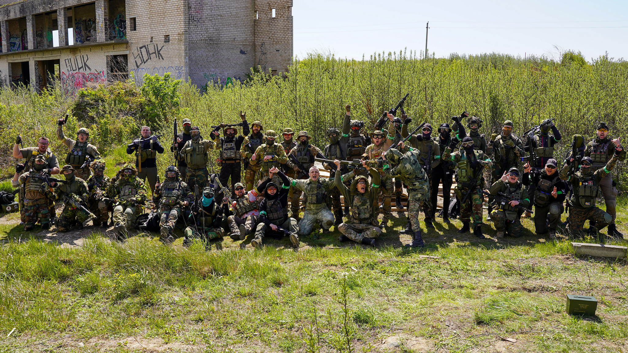 Group of people in military gear posing outdoors in front of overgrown area, with some graffiti on a building in the background.