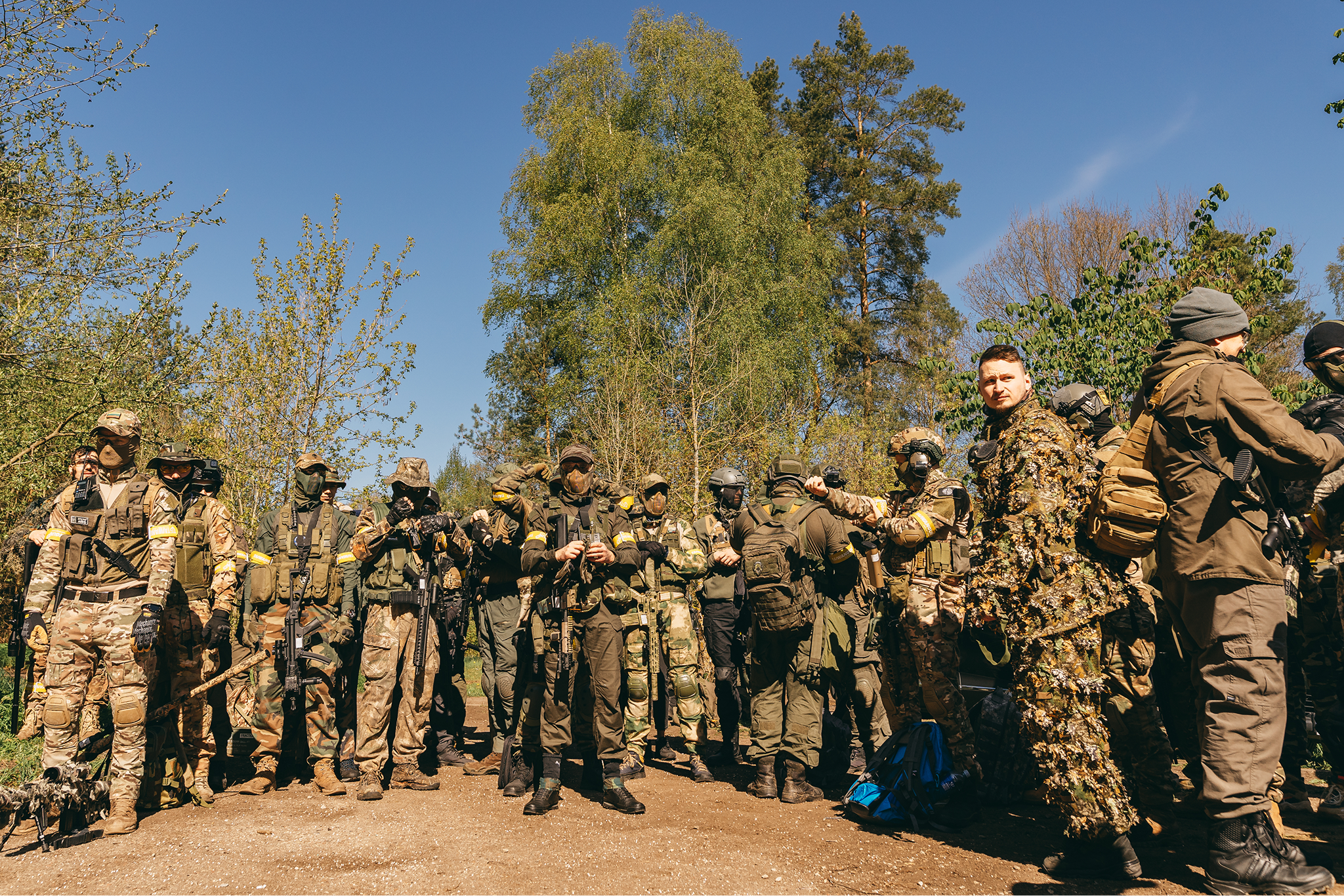 Group of soldiers in camouflage uniforms and tactical gear standing outdoors on a dirt path surrounded by trees under a clear blue sky.