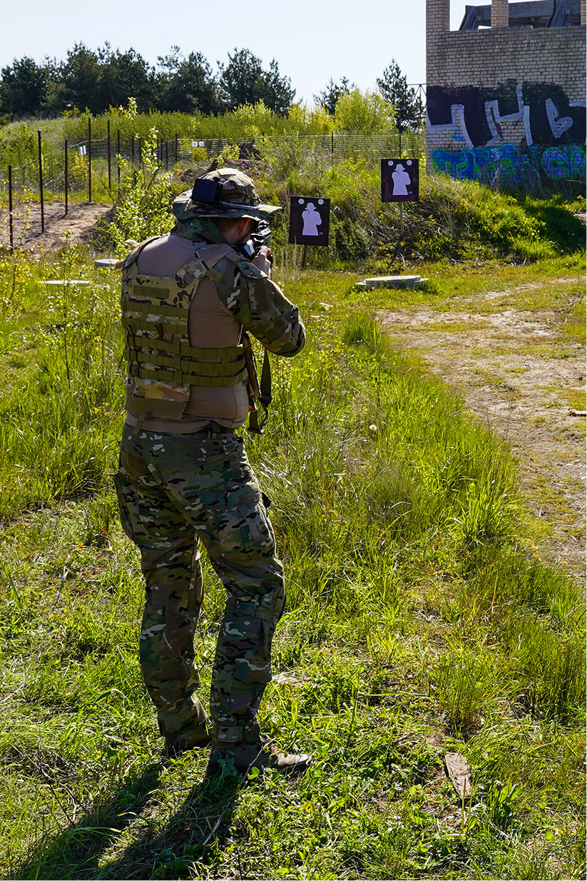 A person in camouflage military attire and a helmet is aiming a rifle at shooting targets in an outdoor practice area with graffiti-covered wall and greenery.