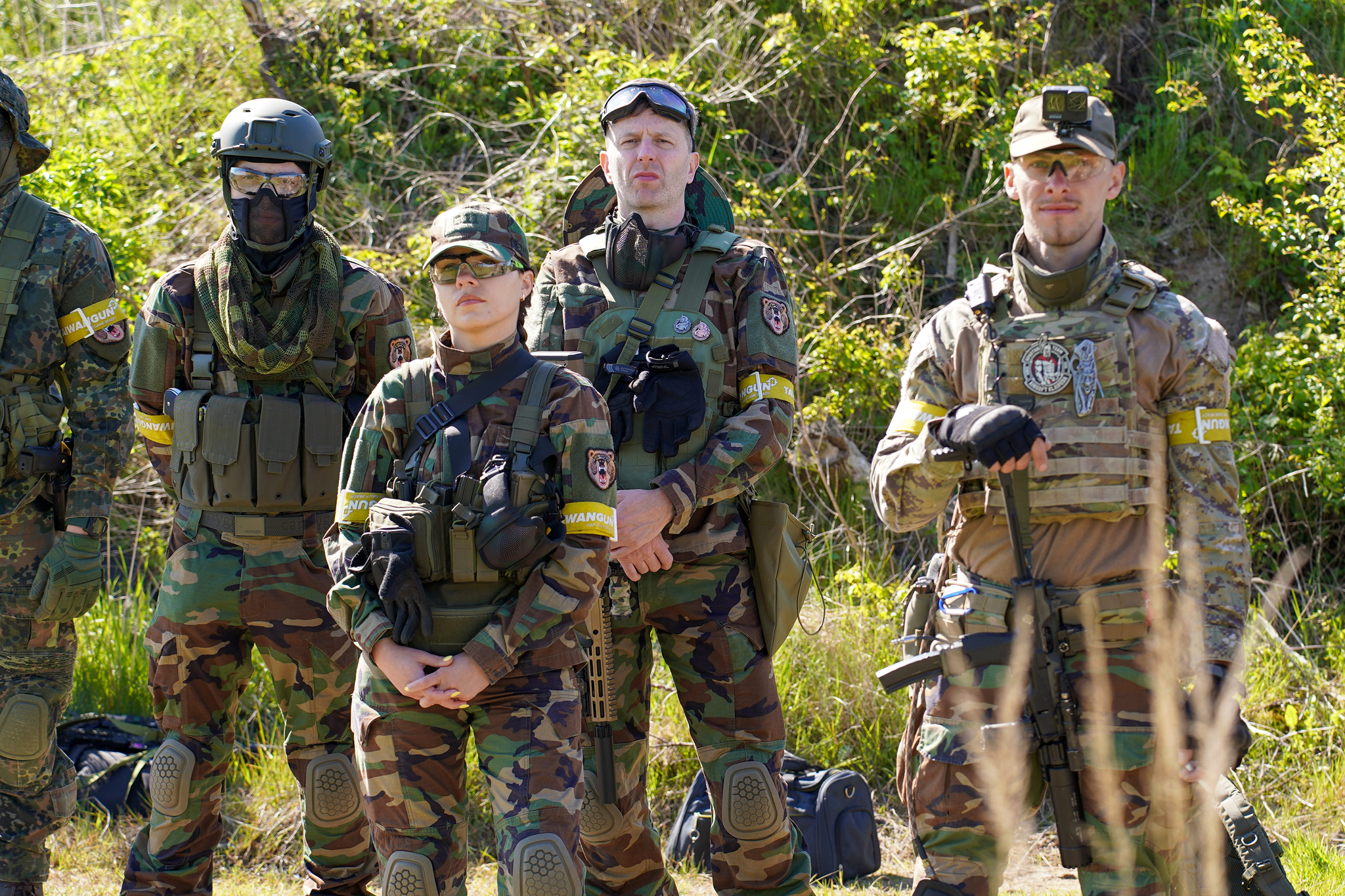 Group of soldiers in camouflage military gear standing outdoors with greenery in the background.