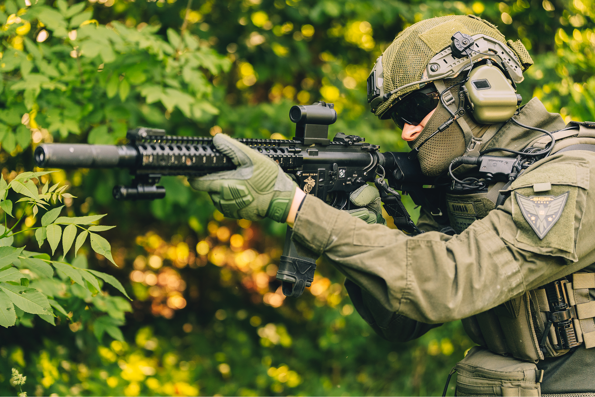 Military personnel in camouflage uniform aiming a rifle in a wooded area.