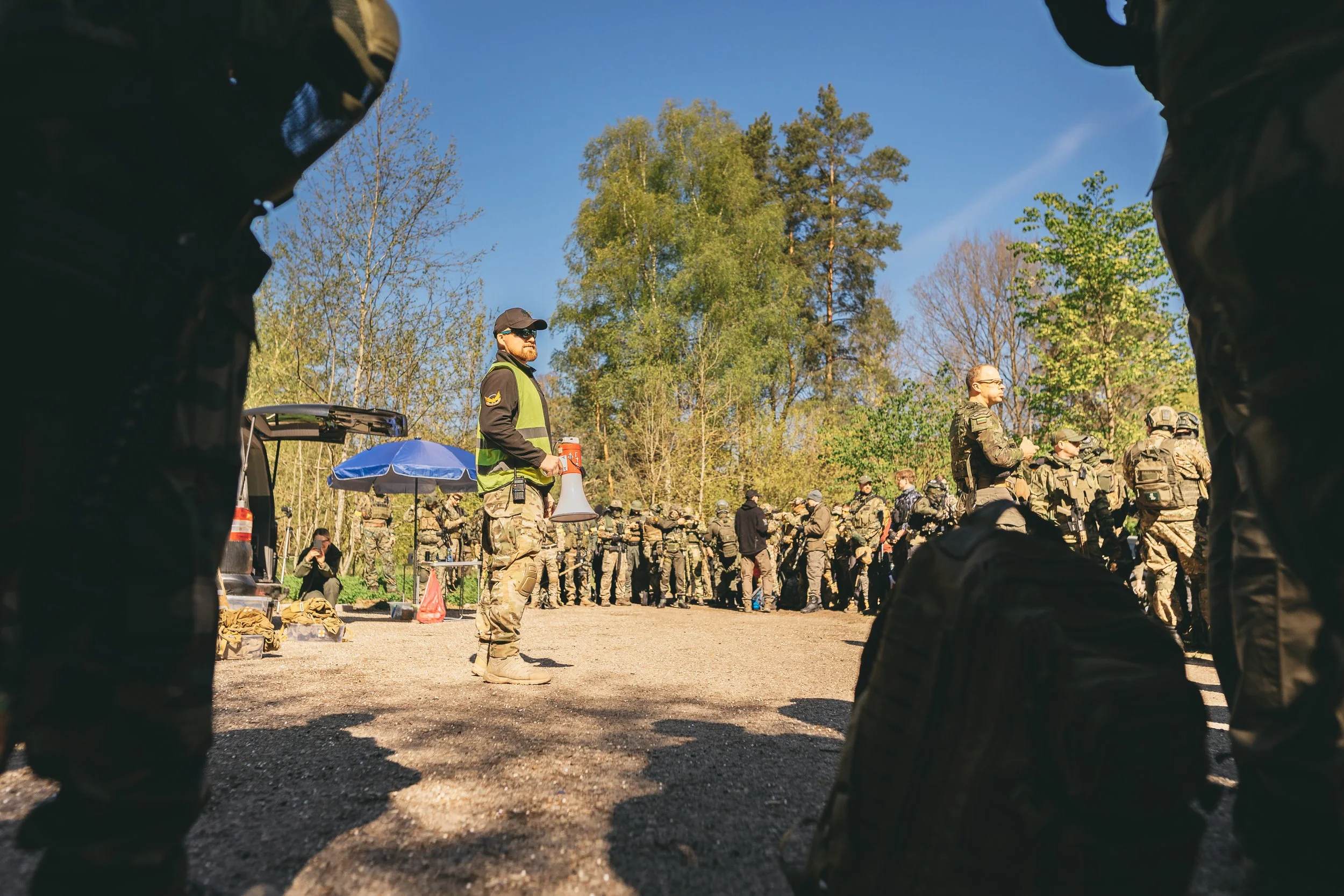 A group of soldiers and military personnel gathered outdoors on a sunny day with green trees in the background.