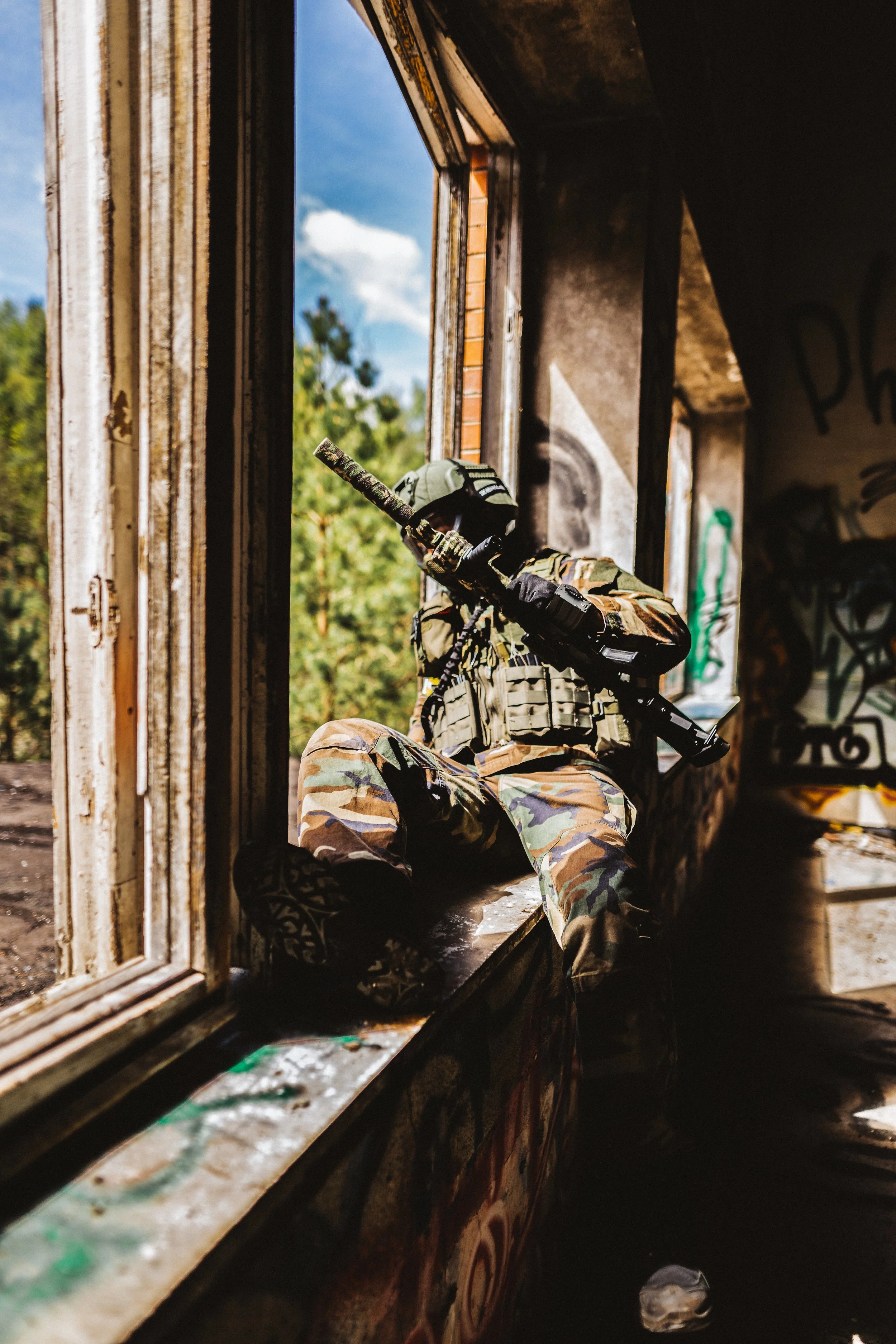 Soldier in camouflage uniform kneeling by a window with a rifle, inside an abandoned building with graffiti on the walls.