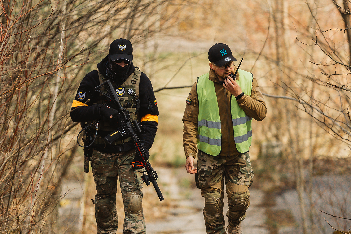 Two armed military personnel walking through a wooded area, one holding a rifle and the other using a walkie-talkie, during autumn.
