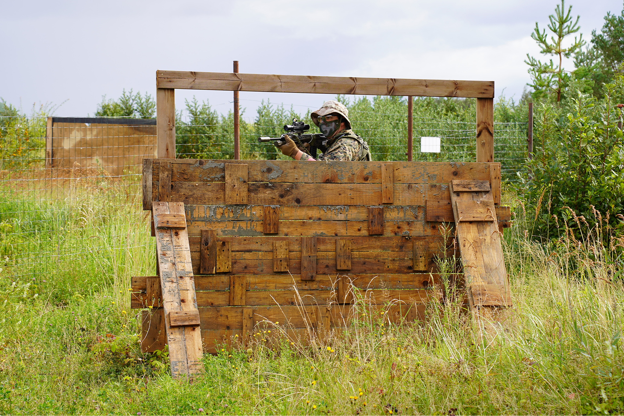 Soldier in camouflage gear and face mask aiming a sniper rifle from a wooden observation post on a grassy field with greenery and trees in the background.