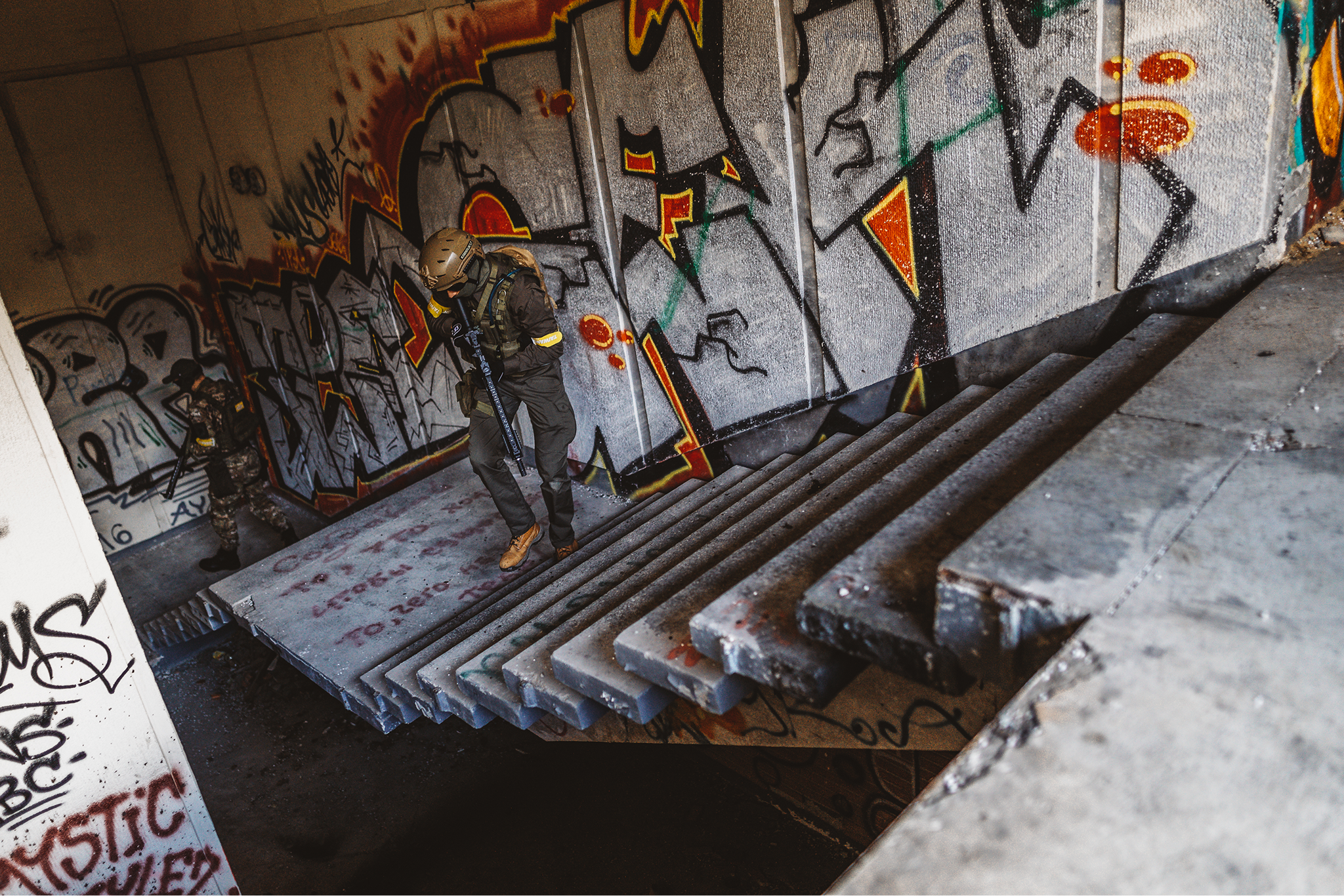 Two armed soldiers in tactical gear descending graffiti-covered concrete stairs in an abandoned building.
