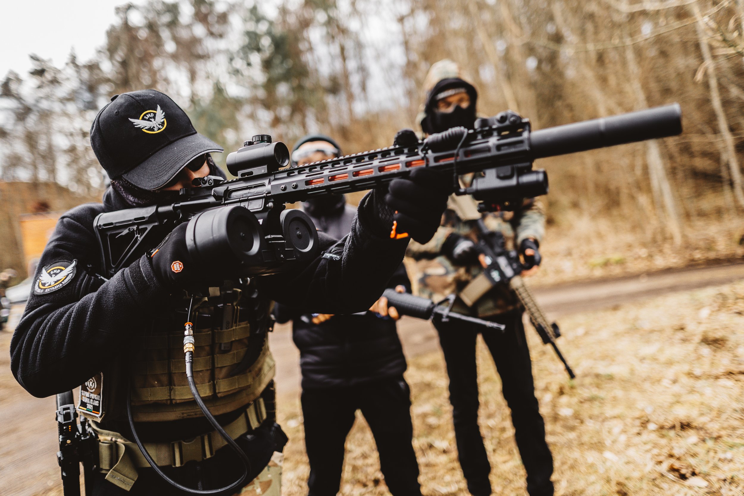 A person in tactical gear aims a large sniper rifle with a scope during a firearm training exercise outdoors, with two other individuals dressed similarly in the background.
