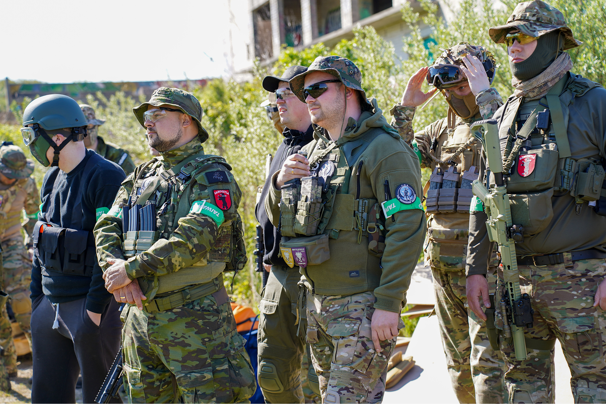 Group of soldiers in camouflage uniforms, some with tactical gear, standing outdoors during daytime with a building and greenery in the background.