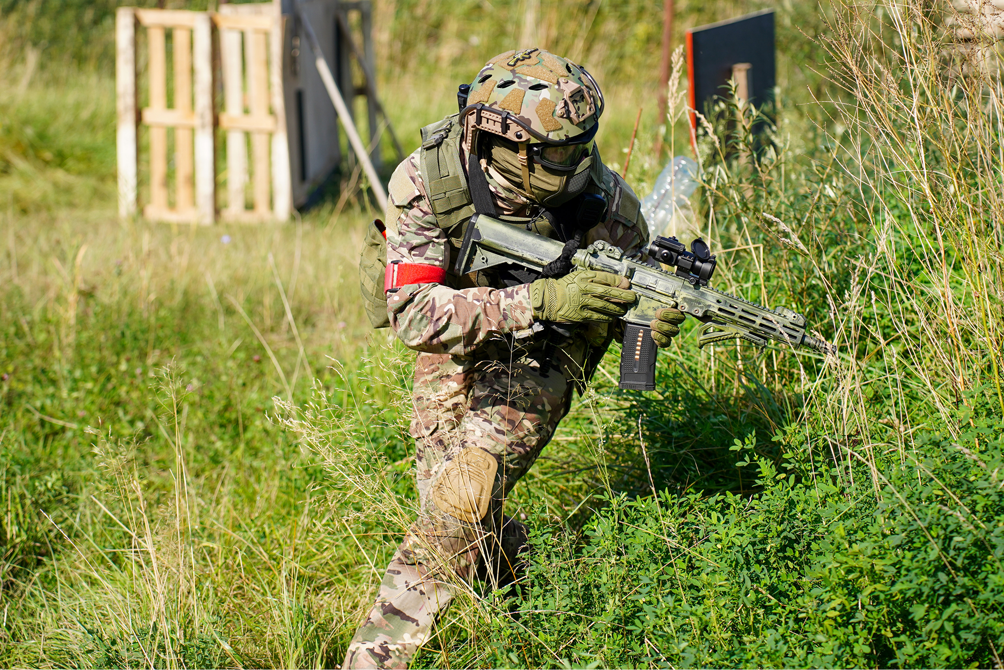 Militarized person in camouflage uniform, tactical gear, helmet, goggles, and gloves, crouching with a rifle in a grassy outdoor environment.