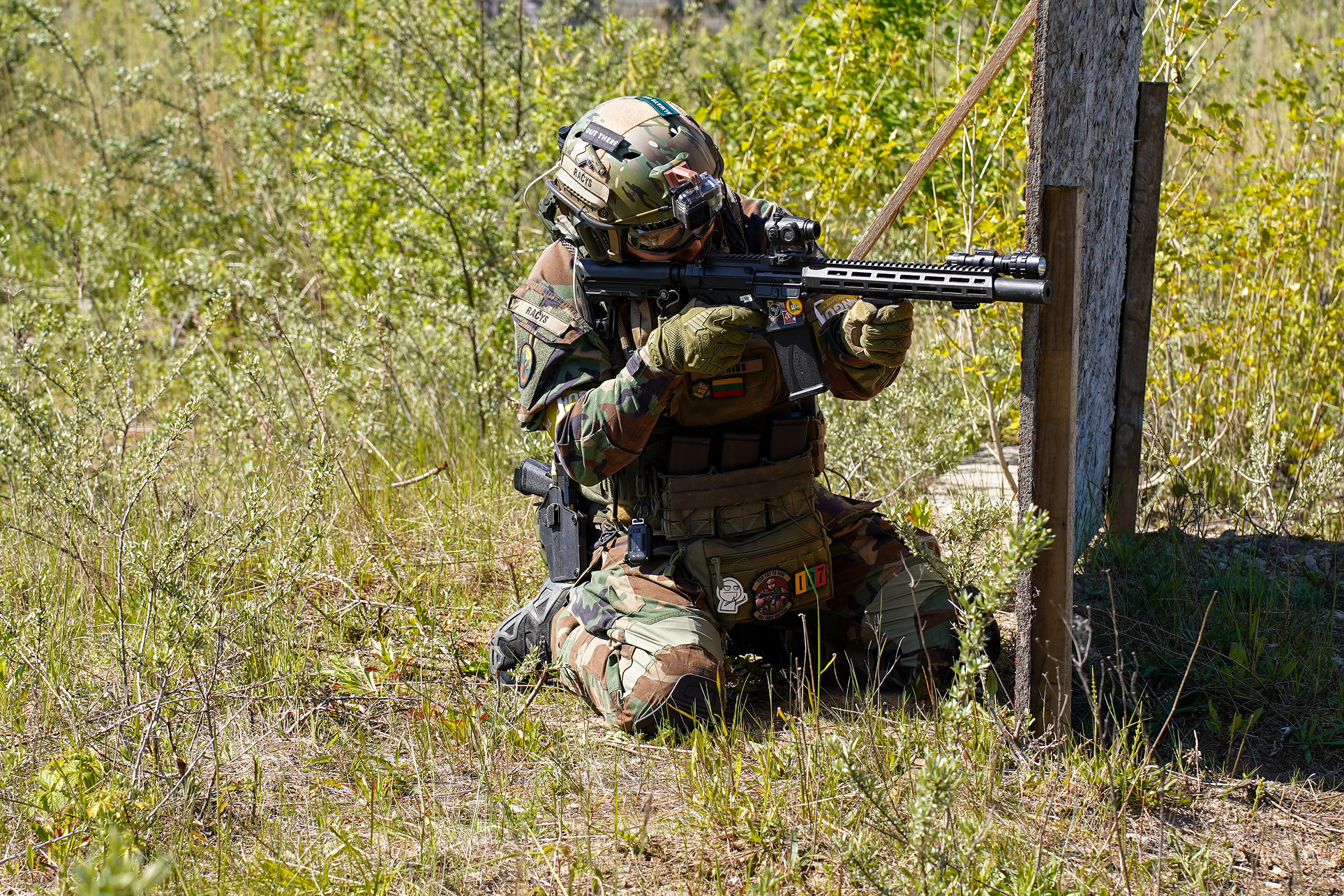 A soldier in camouflage uniform kneels on the ground, aiming a rifle while taking cover behind a wooden post in a grassy field.