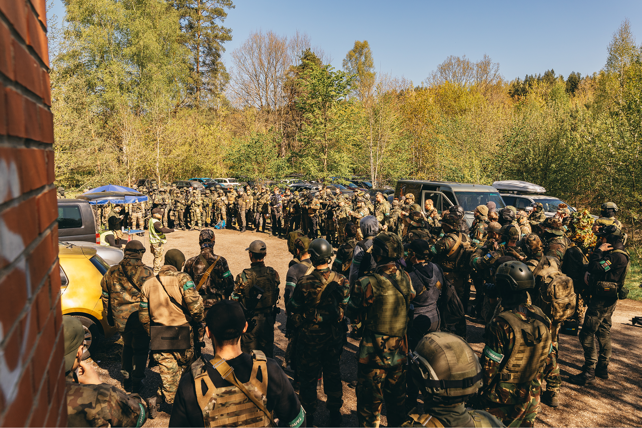 A large group of armed soldiers in camouflage military uniforms gathered outdoors, with trees and parked cars in the background during daytime.