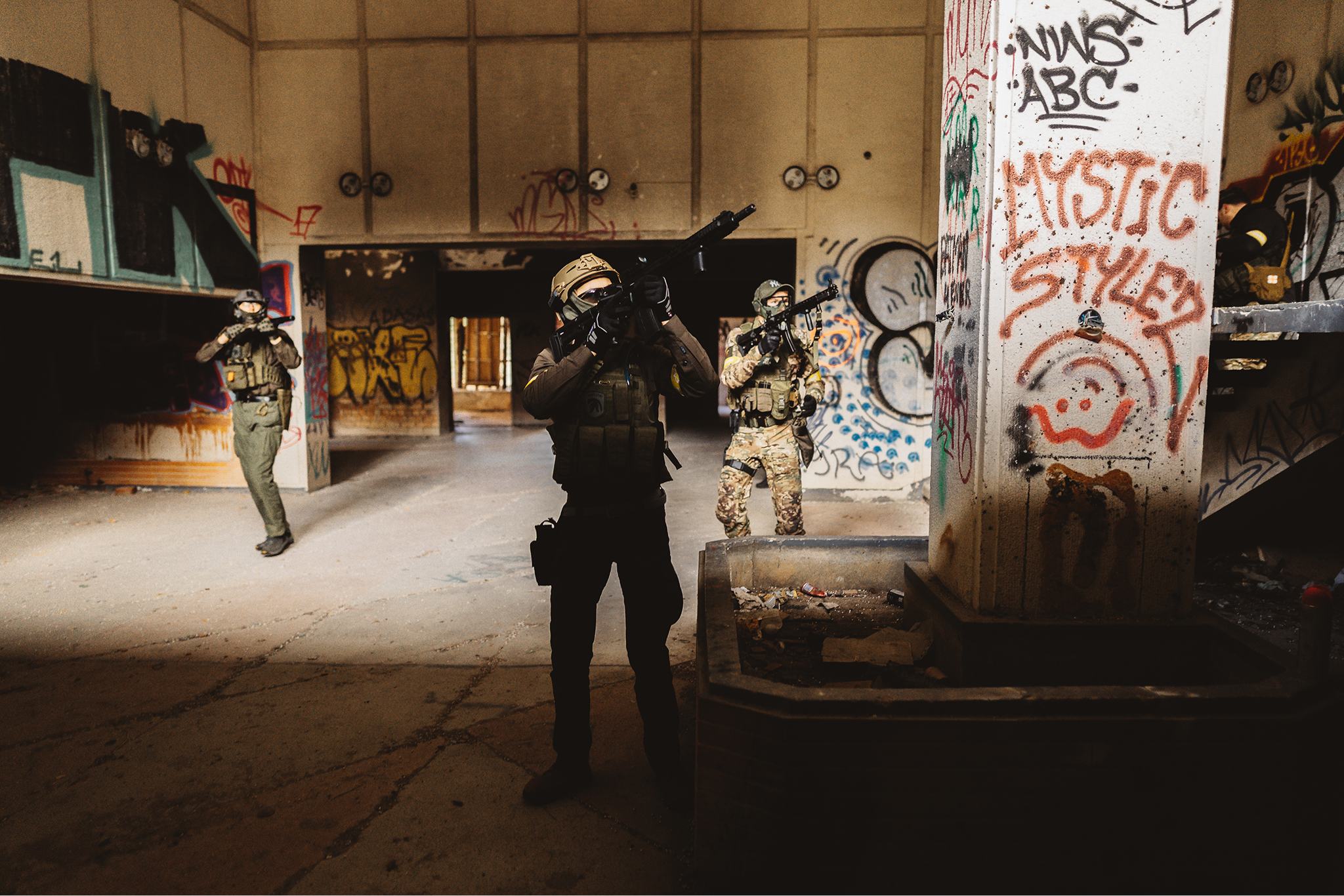 Three armed individuals wearing tactical gear and camouflage clothing inside an abandoned, graffiti-covered building.