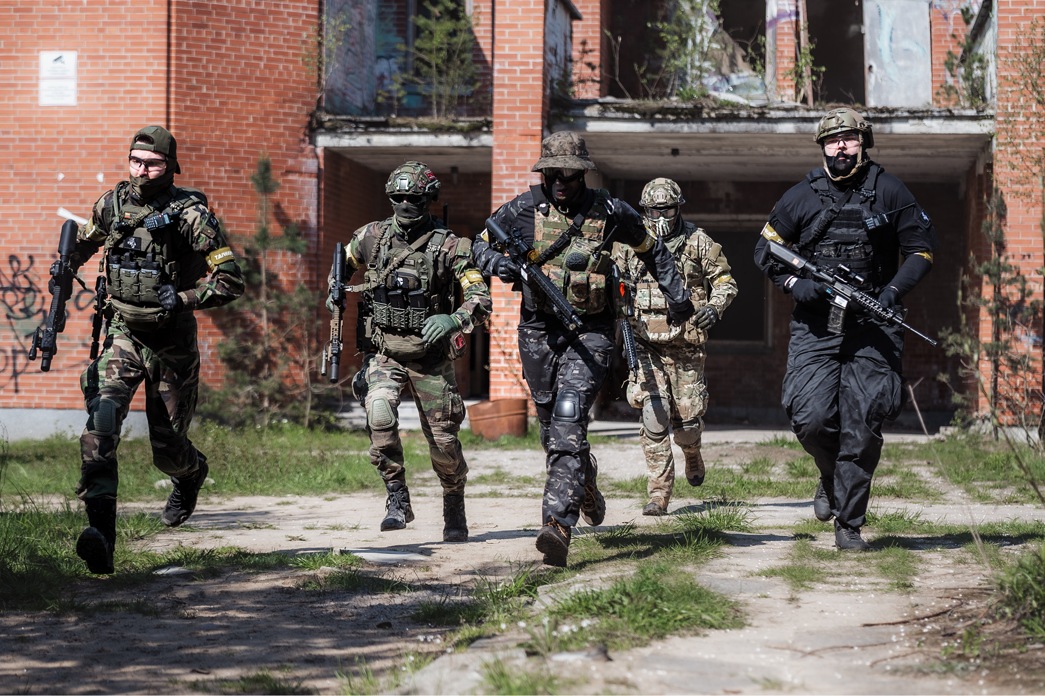 Military or tactical personnel running in front of a damaged brick building.