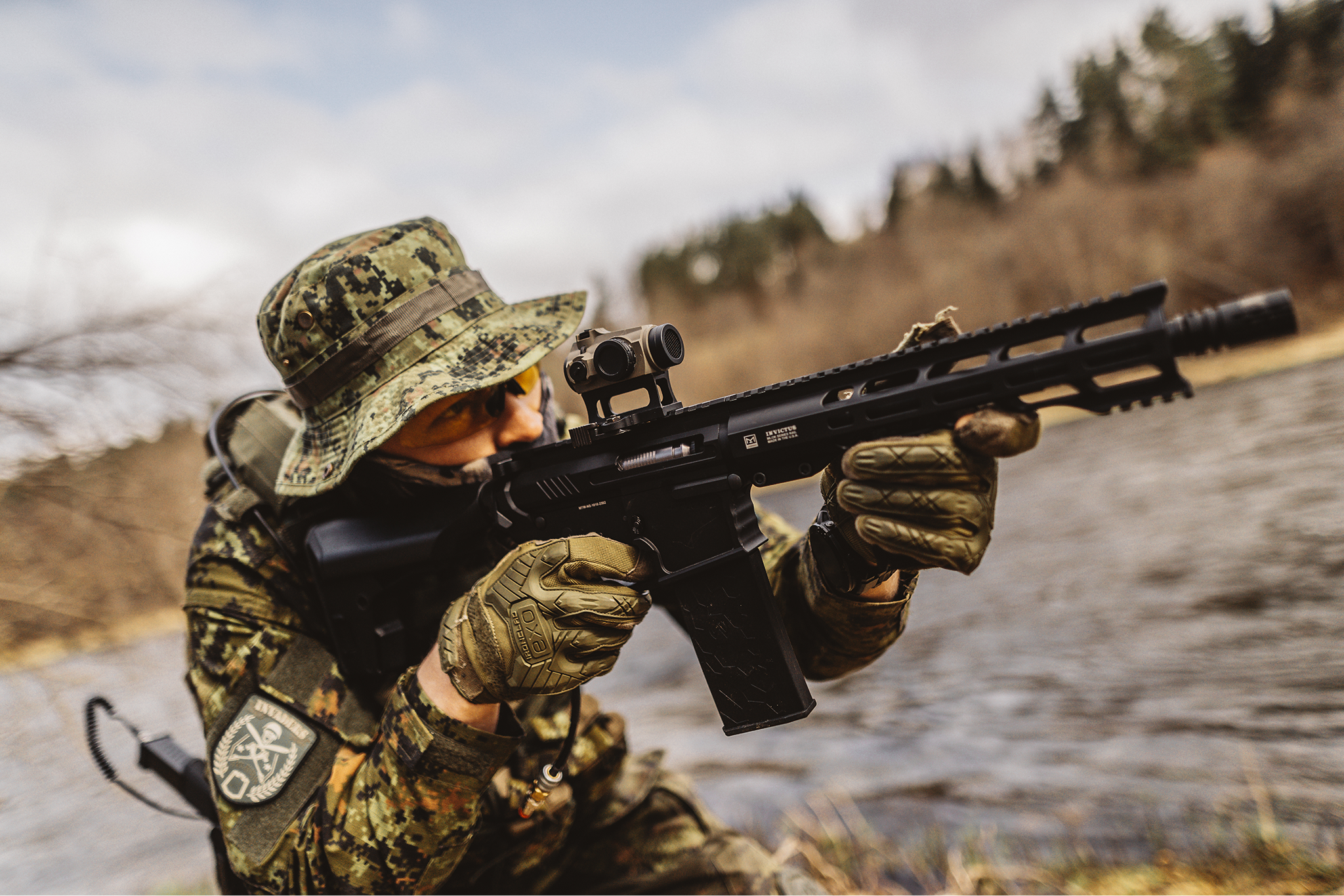 Soldier in camouflage gear aiming a black rifle with a scope while crouching outdoors near a river with trees in the background.