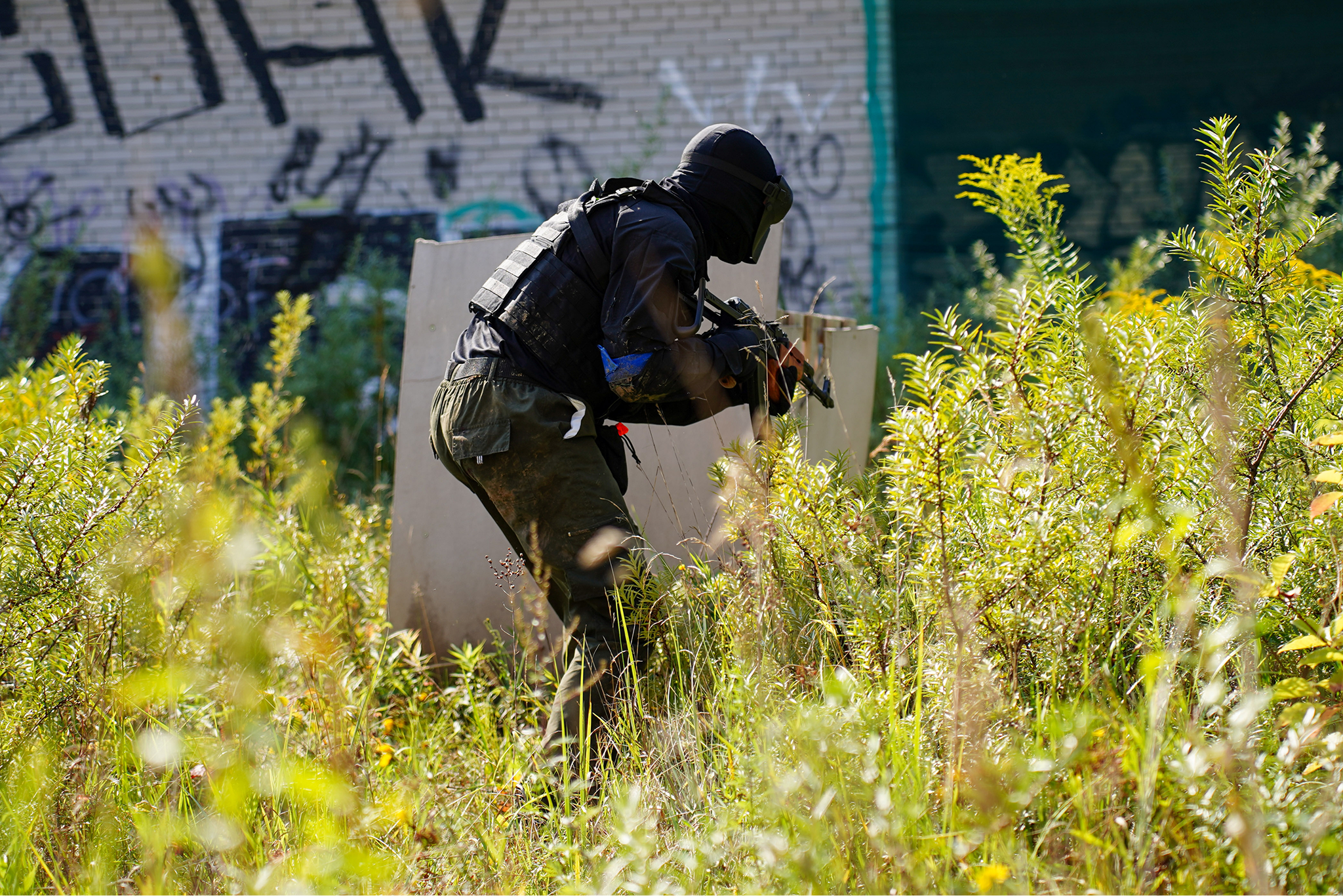 A person dressed in tactical gear and a helmet, crouching behind a shield and holding a weapon, in a grassy outdoor area with graffiti-covered walls in the background.