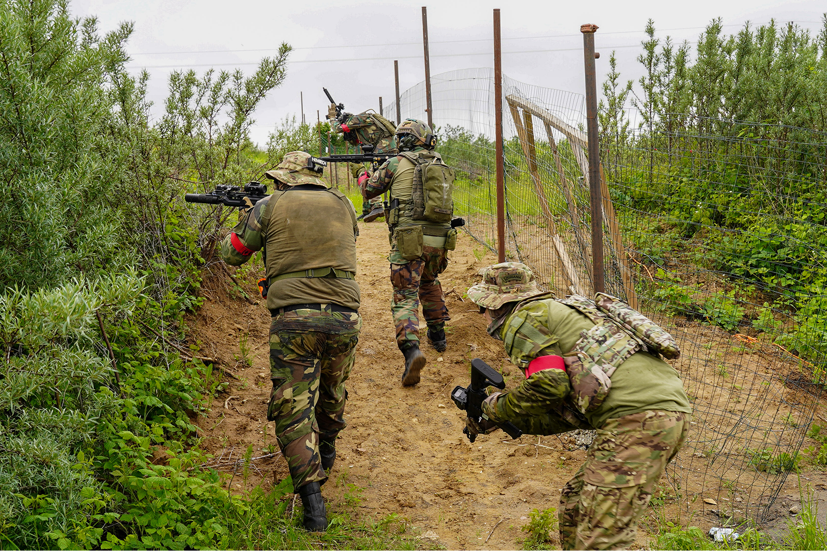 Four soldiers in camouflage uniforms and gear crouch and move along a dirt path bordered by greenery and a wire fence, during a military training exercise.