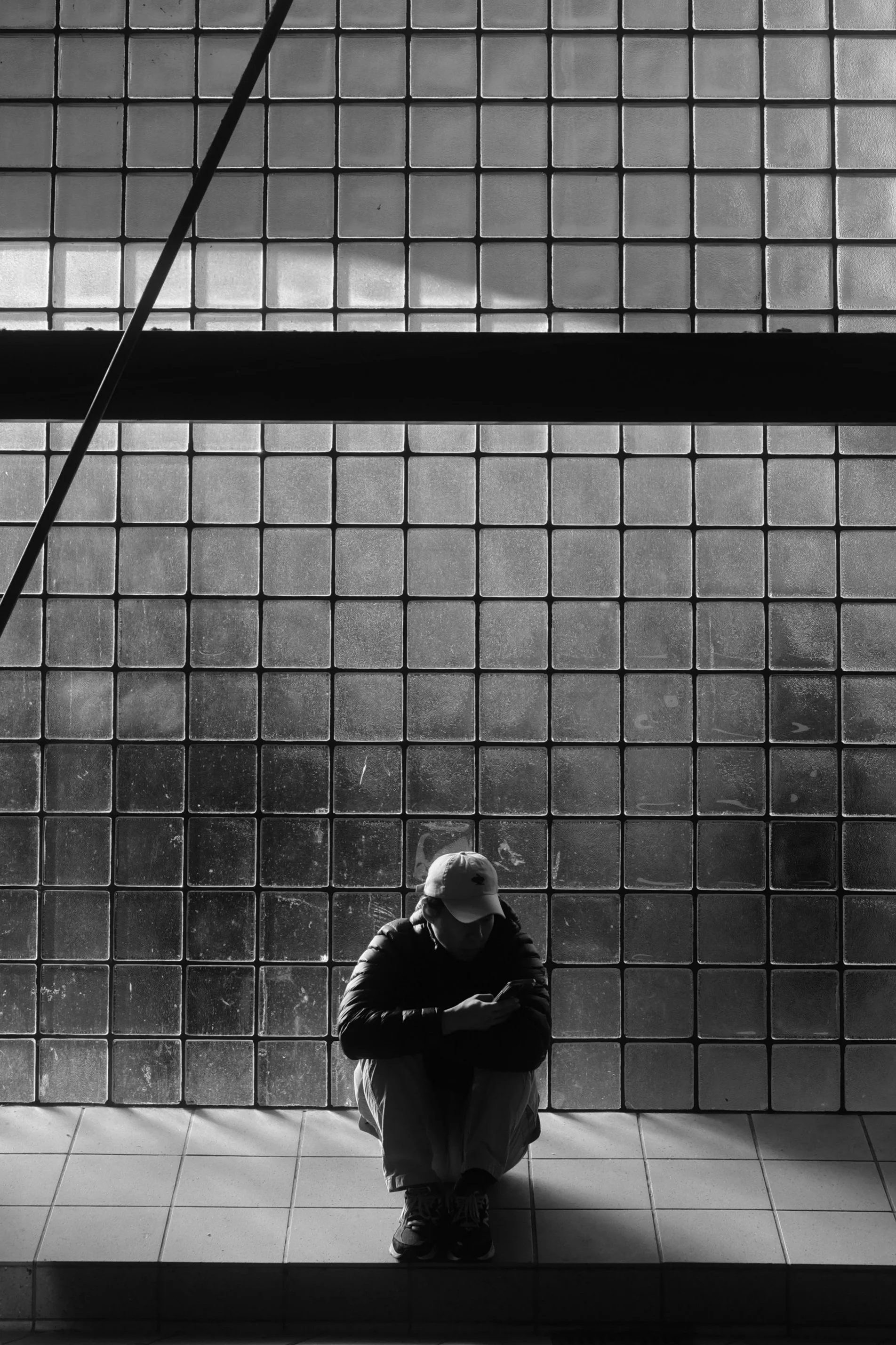 Black and white photo of a person wearing a cap, squatting on a tiled floor, with a backdrop of glass block windows.