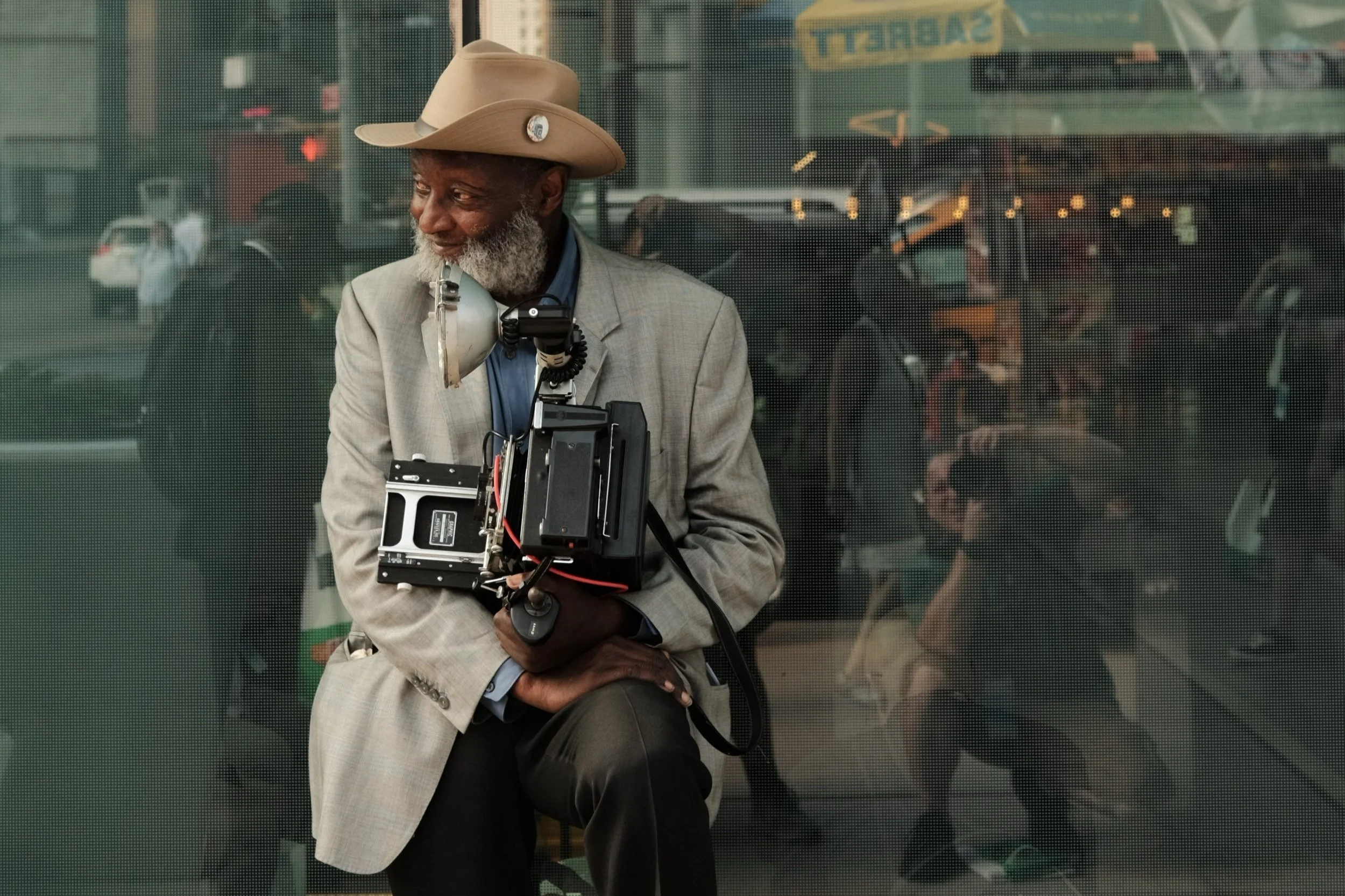 An elderly man wearing a cowboy hat and suit jacket is holding a vintage camera. He's sitting in front of a glass window that reflects city street scenes and people, including a photographer capturing the shot.