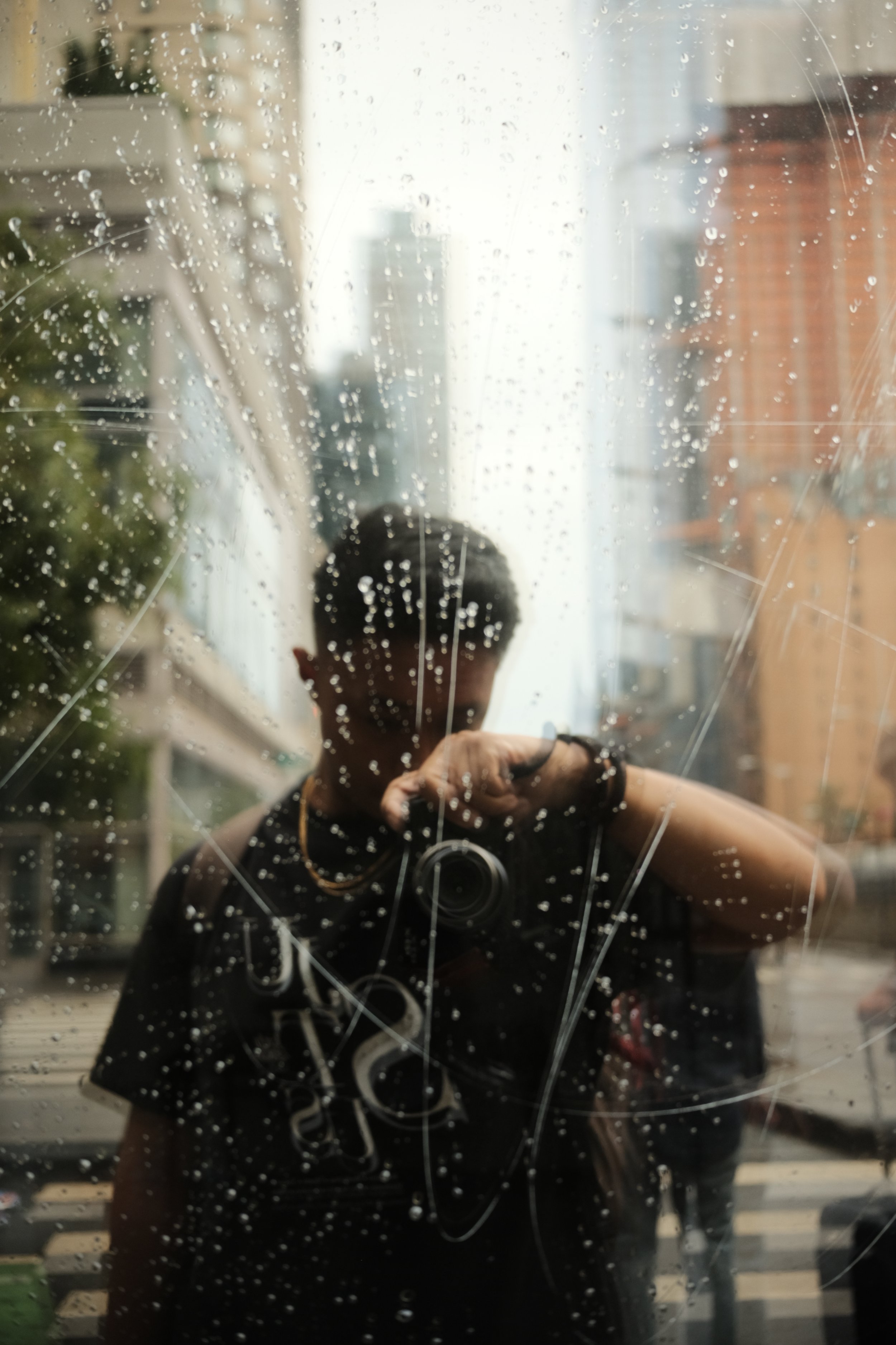 Person taking a photo through a window with raindrops, capturing urban street reflections.