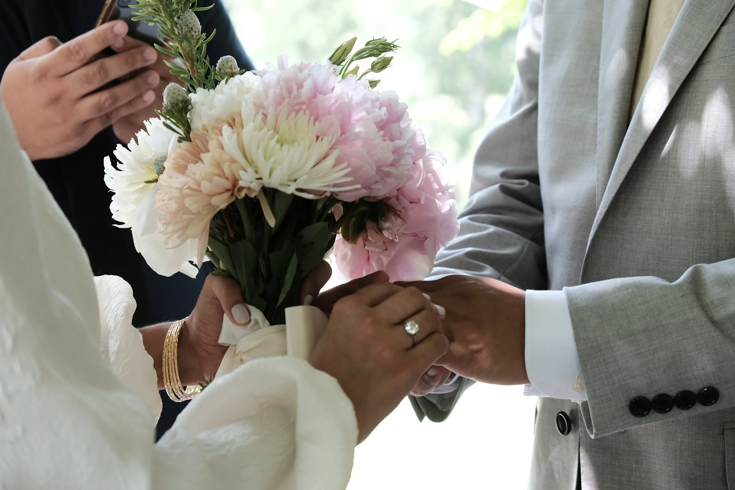 Close-up of a couple exchanging rings during their wedding ceremony, with a bouquet of pink and white flowers.