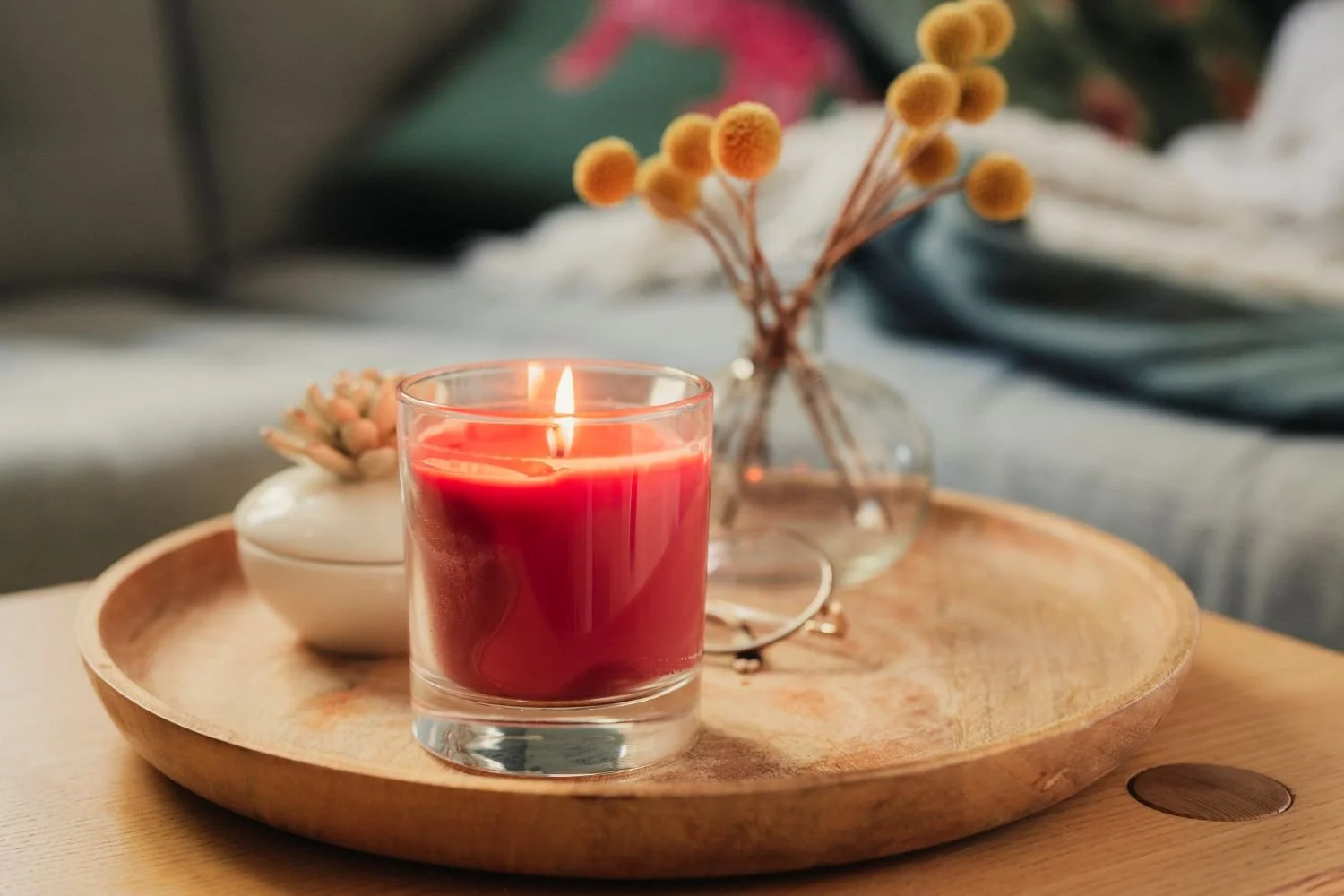 Red candle, light flowers, and a white ceramic bowl on a wooden tray.