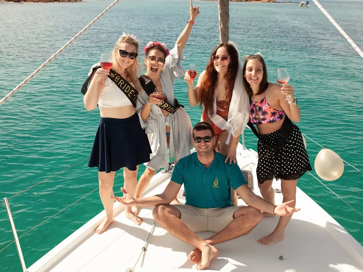 Group of five friends celebrating on a boat in the water, some wearing 'bride' sashes, with glasses of rosé wine, during a bachelorette party.