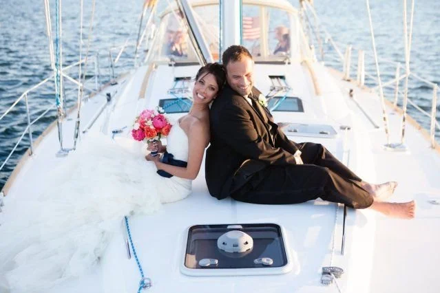 A newlywed couple sitting on the deck of a sailboat, smiling. The bride holds a bouquet of pink flowers, and the groom is barefoot, wearing a black suit.