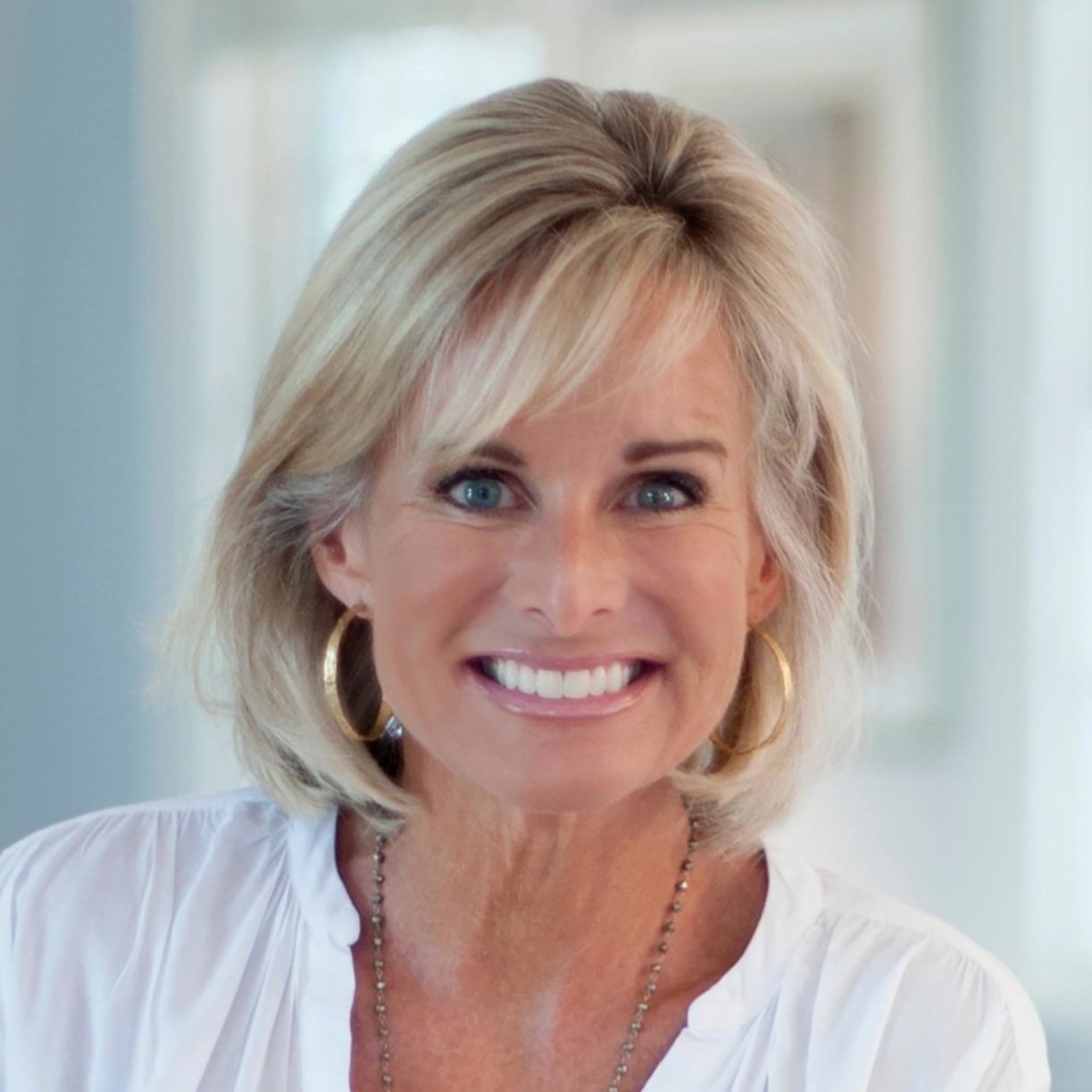 A smiling woman with blonde hair, blue eyes, wearing gold hoop earrings, a white top, and a necklace.