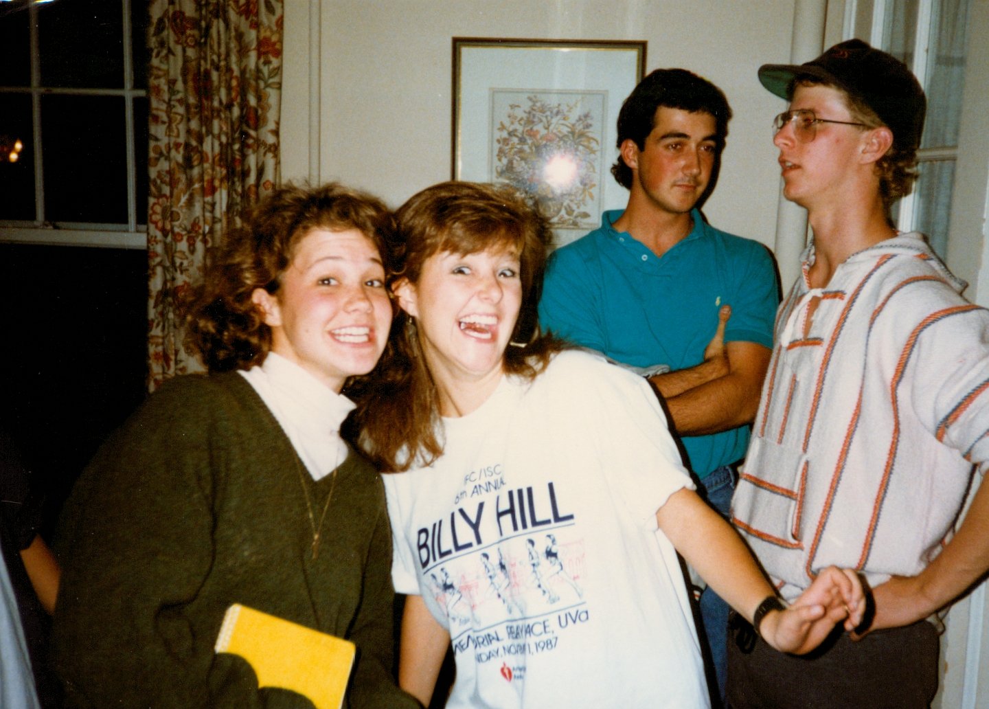 Four young adults, two women and two men, are standing indoors, smiling and posing for a photo. The women are on the left, with one holding a yellow book, and the men are on the right, one with crossed arms and the other wearing glasses and a cap. Th