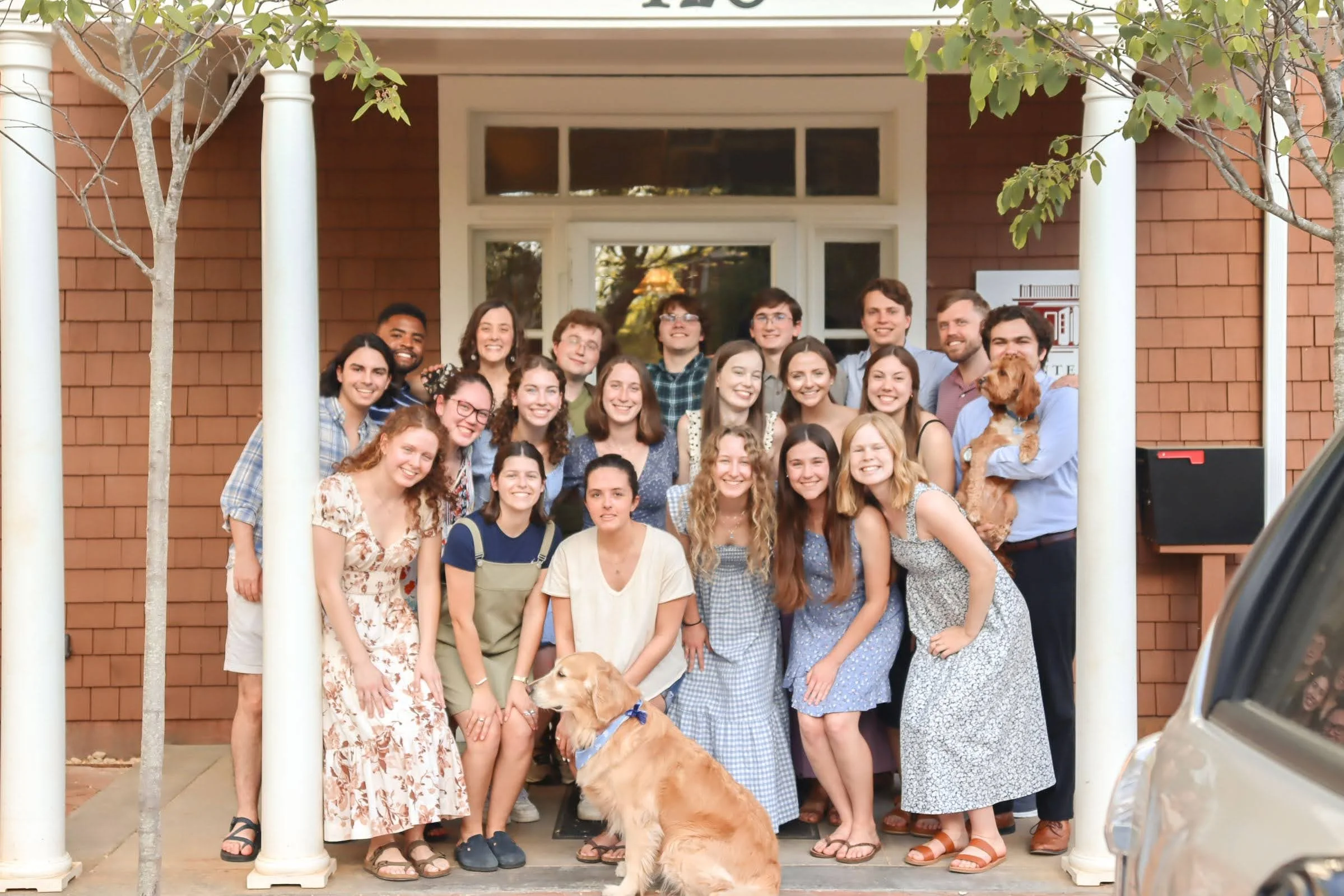 A group of young people gathered on the porch of a brick house, posing for a photo with two dogs, one sitting on the ground and one being held by a person. The group is smiling and appears cheerful.