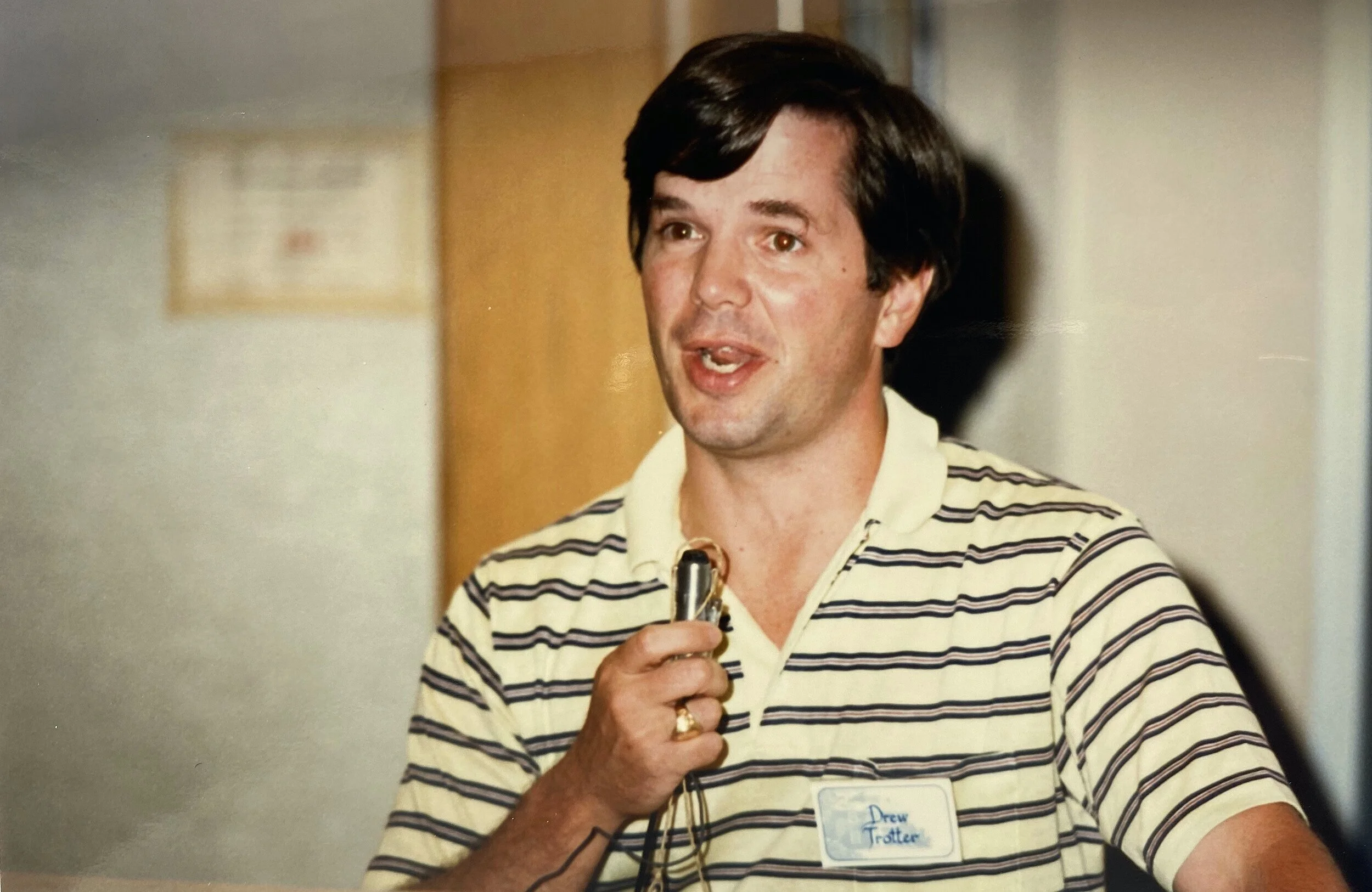 A man with dark brown hair, wearing a striped polo shirt, holding a microphone, speaking at an event. He has a name tag that reads "Drew J Trottier."