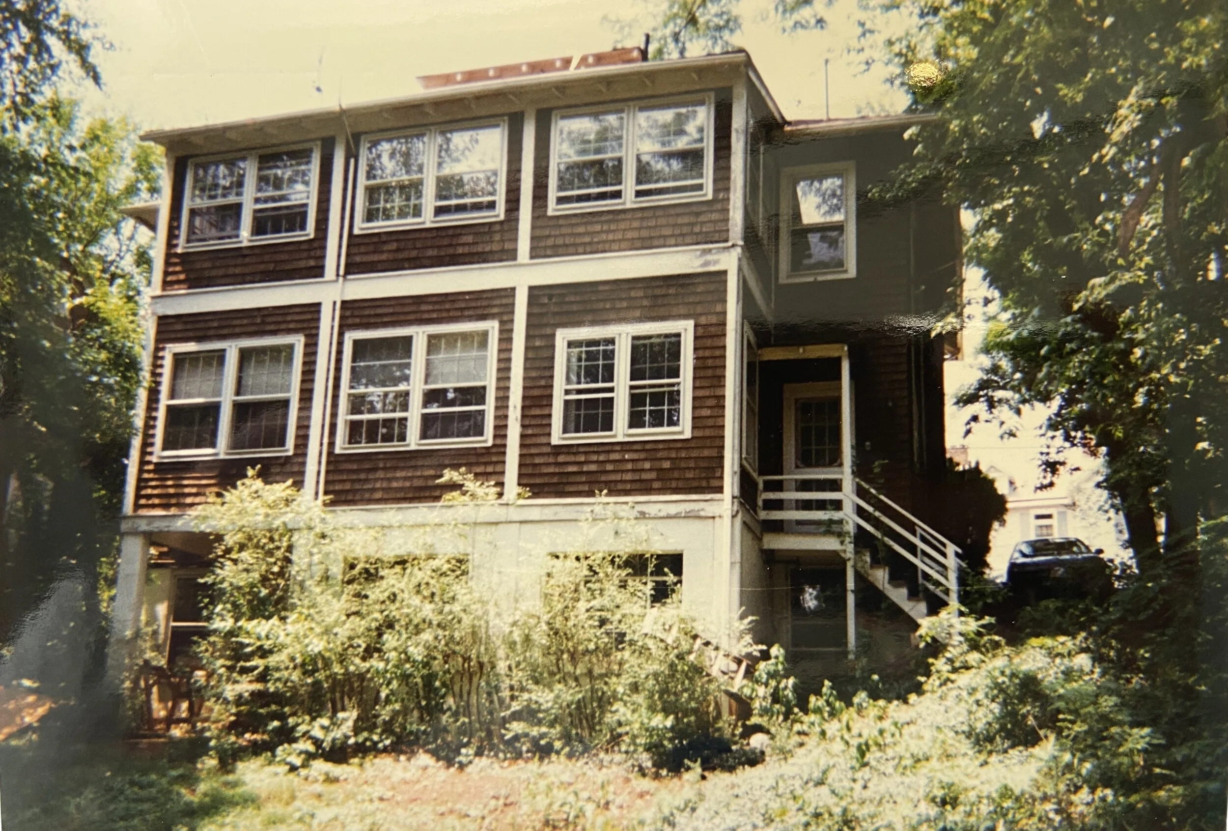 A multi-story house with brown wooden shingles, multiple white-framed windows, a stairway leading to a side entrance, and a car parked on a driveway in the background, surrounded by trees and greenery.