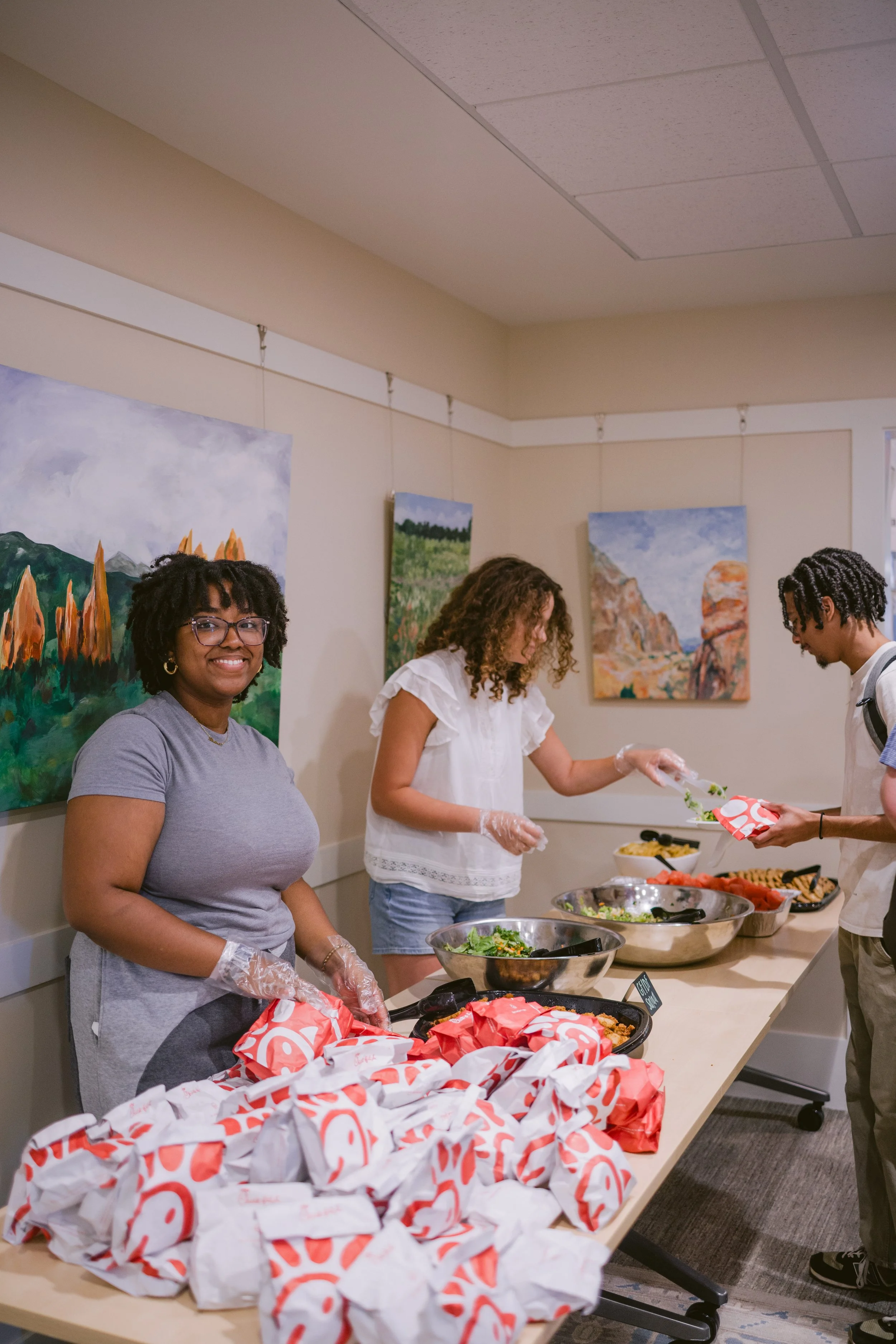 A group of people in a room serving food at a buffet table with Chick-fil-A paper bags. One woman in a gray shirt and glasses is smiling at the camera. Other people are seen preparing or serving food. The room has paintings of landscapes on the wall.