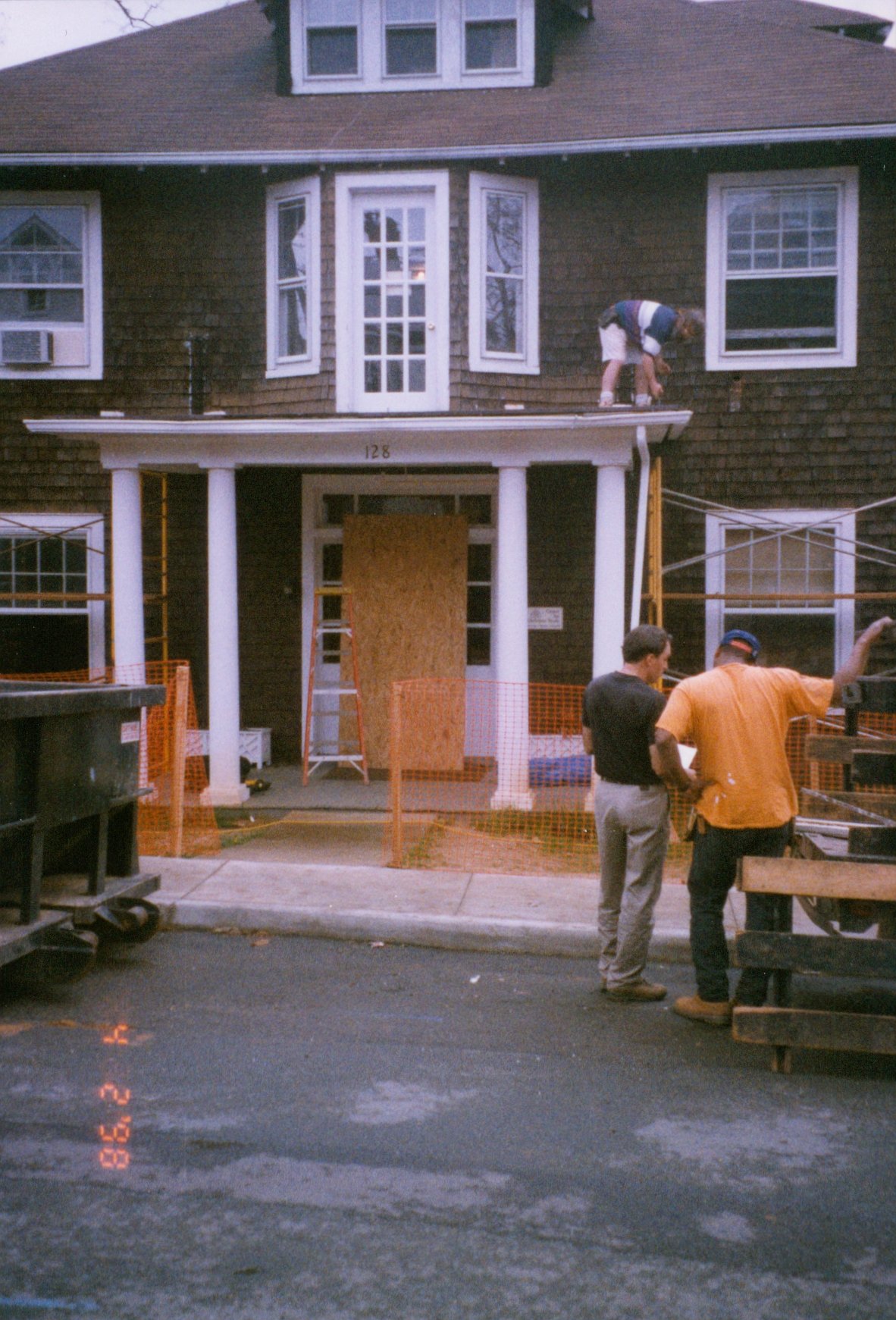 Construction workers working on the front porch of a house, with one person on the roof and two people talking on the ground in front, surrounded by safety fencing.