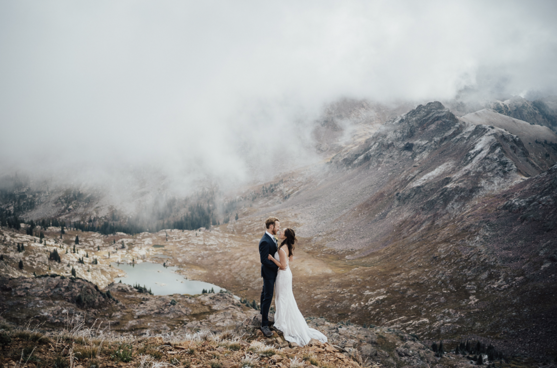 A bride and groom standing on a mountain with a scenic lake and rugged mountains in the background, surrounded by clouds.