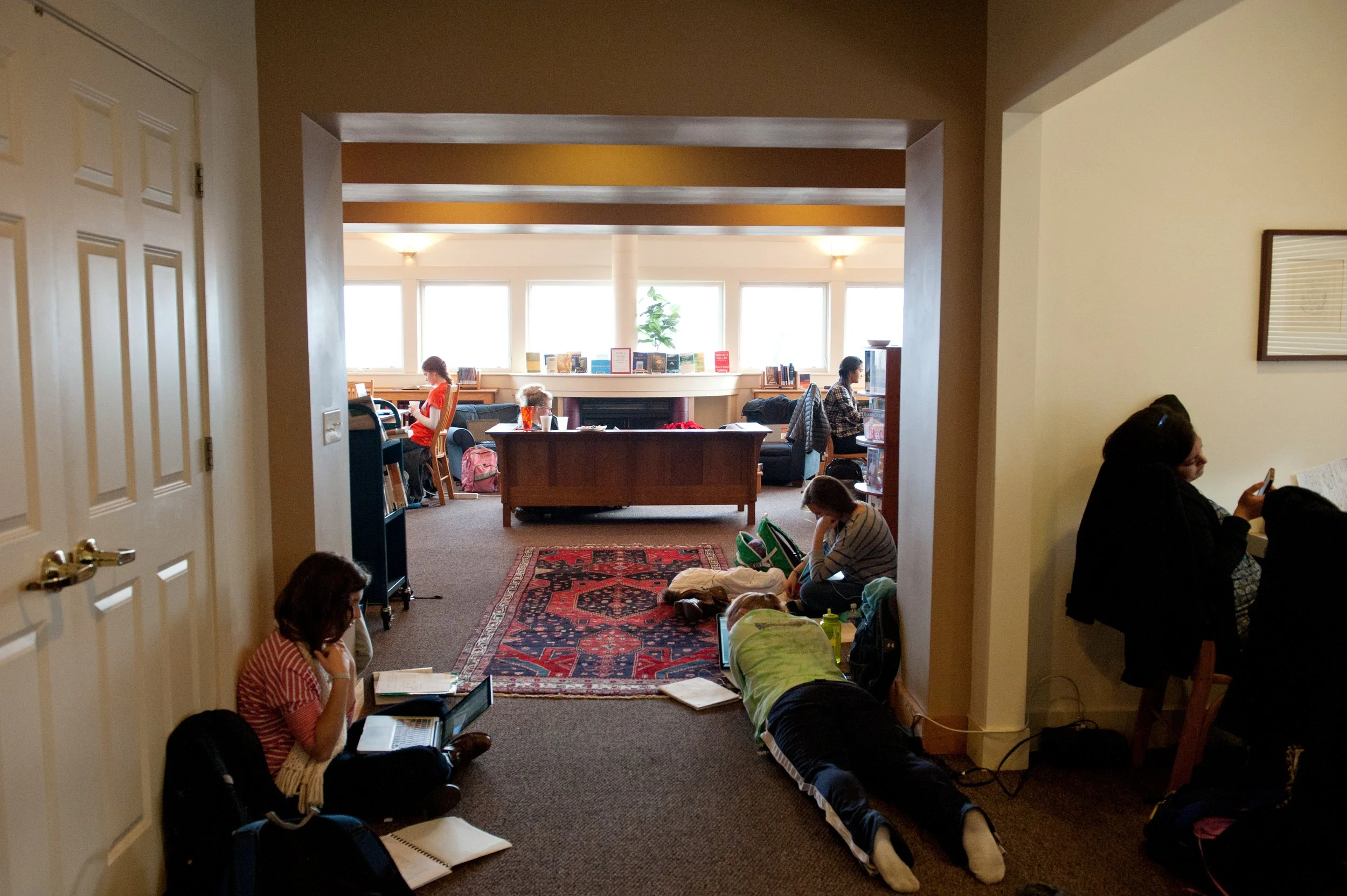 People sitting and reading in a cozy library room with large windows and a fireplace.