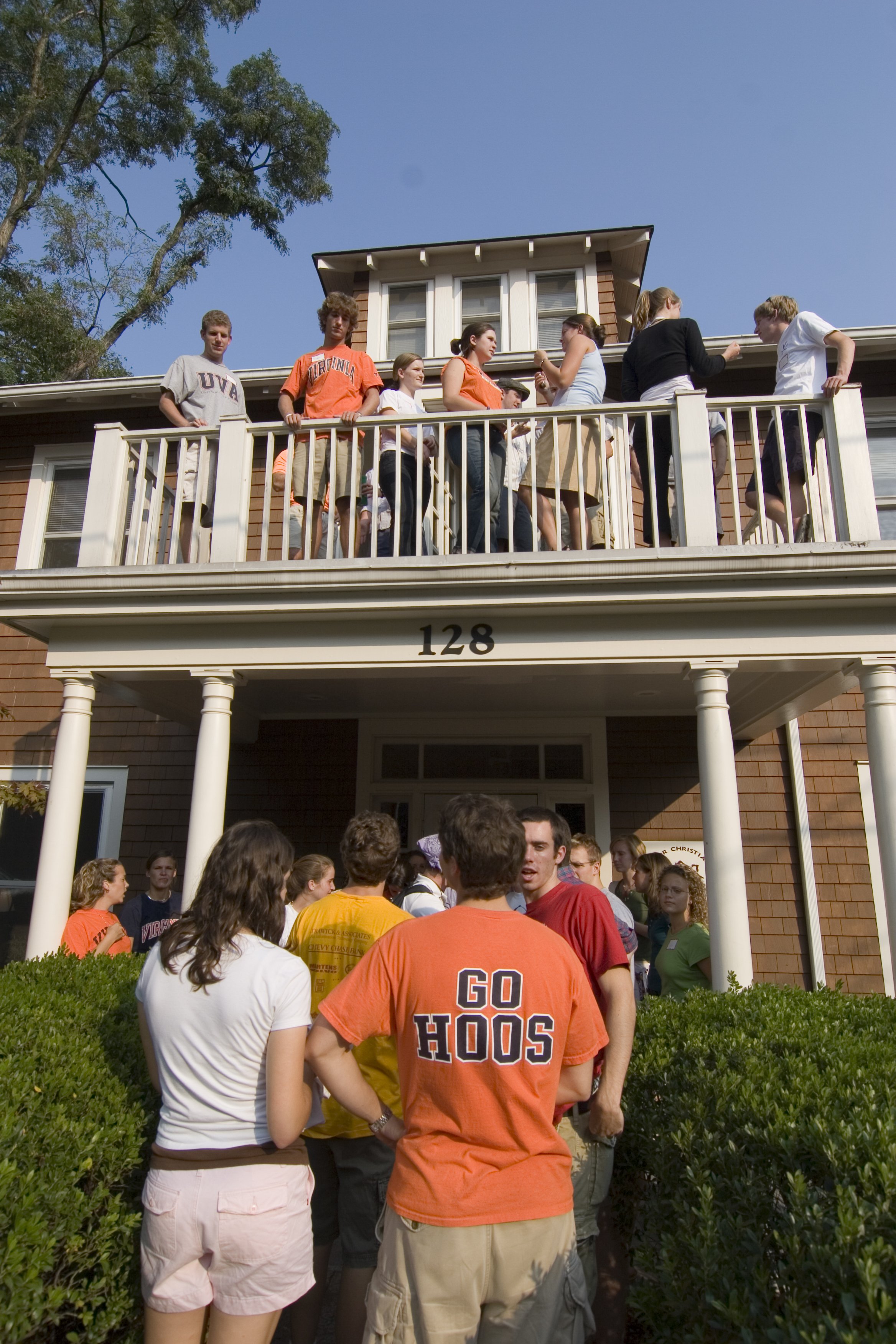 Group of young people gathering outside a house with the number 128 on it, some standing on the porch and others on the lawn, engaging in conversation. Many are wearing casual clothing, and some are wearing shirts with college or team names and sloga