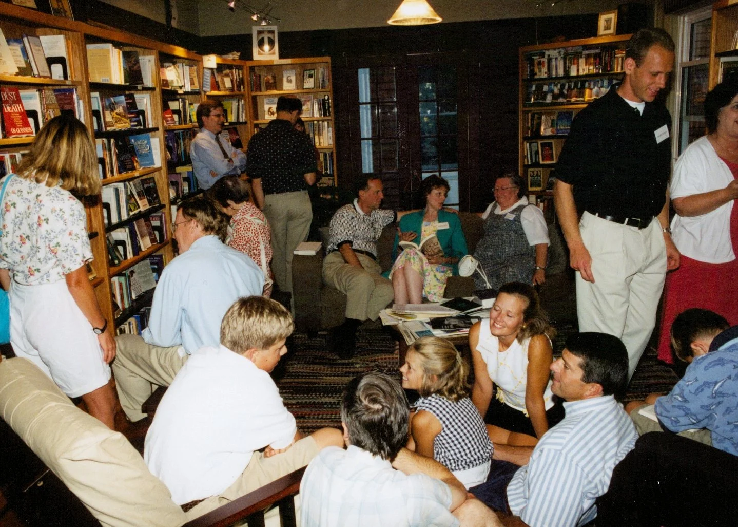 A group of people socializing in a cozy, wood-paneled room filled with bookshelves, some sitting on couches and chairs, others standing and talking, with a table covered in papers and books.